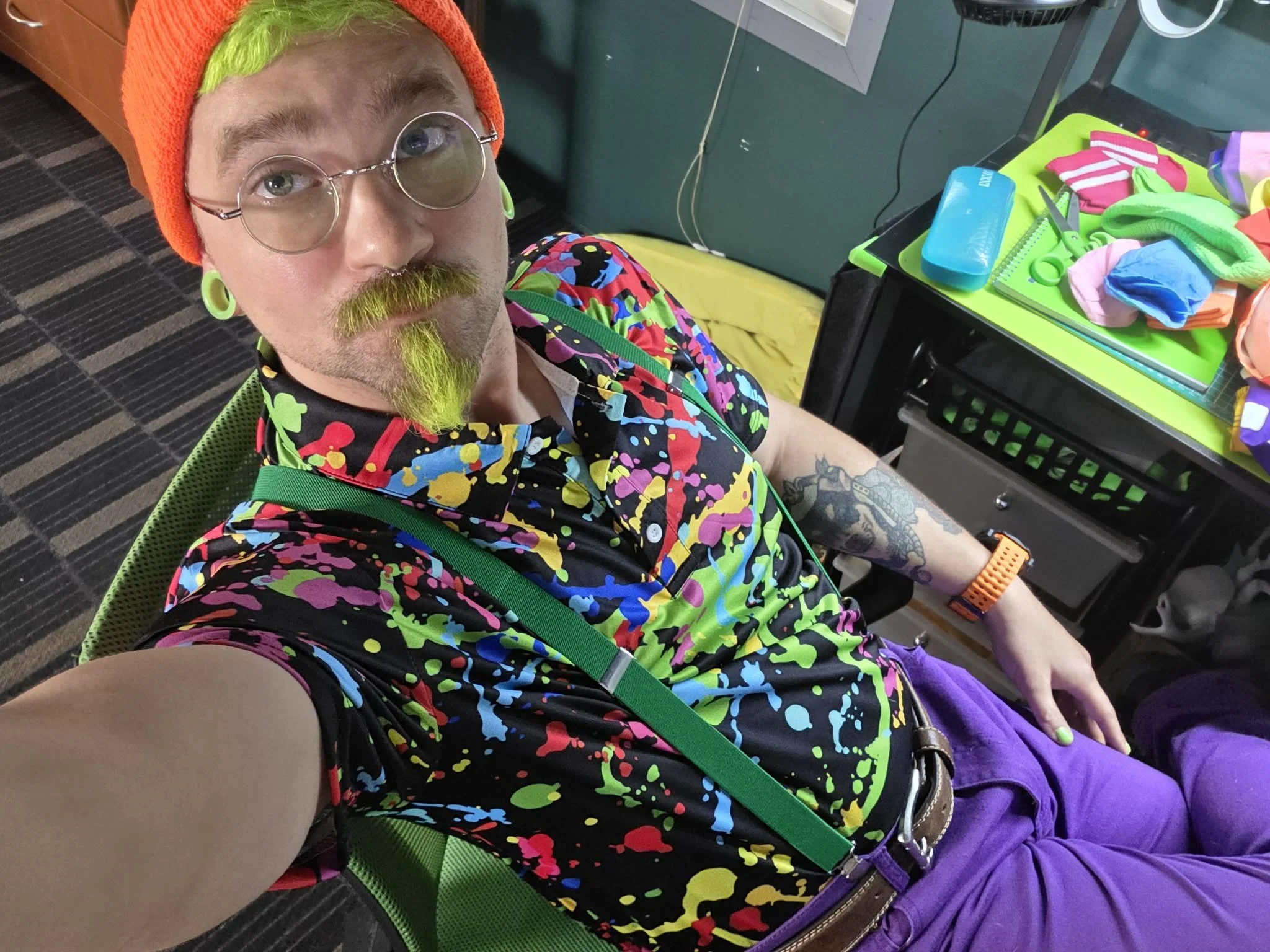 A person with neon green hair, yellow facial hair, and large round glasses taking a selfie in a colorful, patterned shirt, purple pants, and wearing a neon orange beanie and matching earrings. They are sitting at a desk with various colorful items, scissors, and a tape measure.