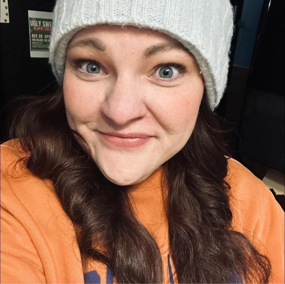 A woman with long brown hair, wearing a white knit beanie and an orange top, smiling at the camera.