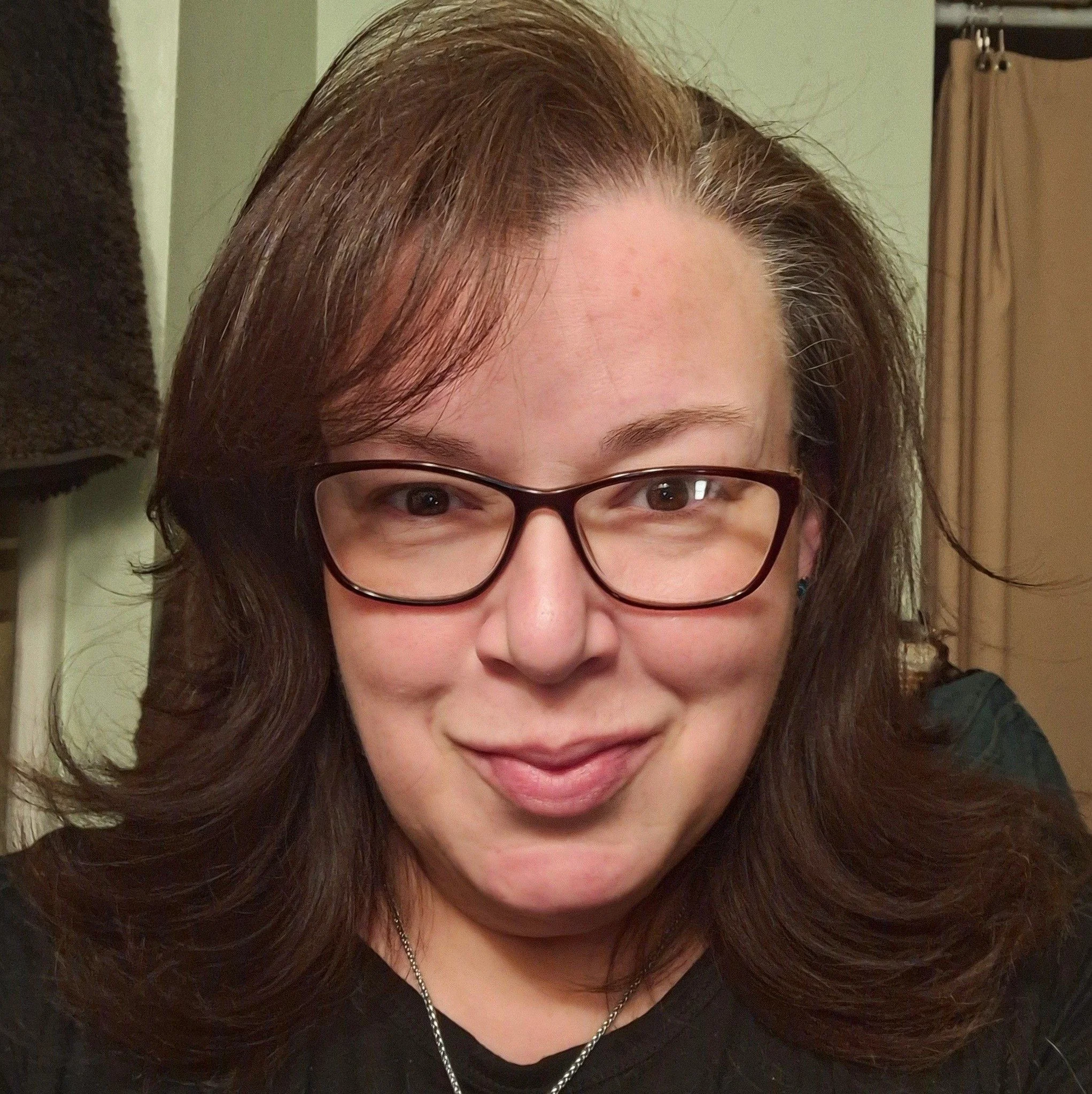 A woman with glasses and long brown hair smiling at the camera indoors.