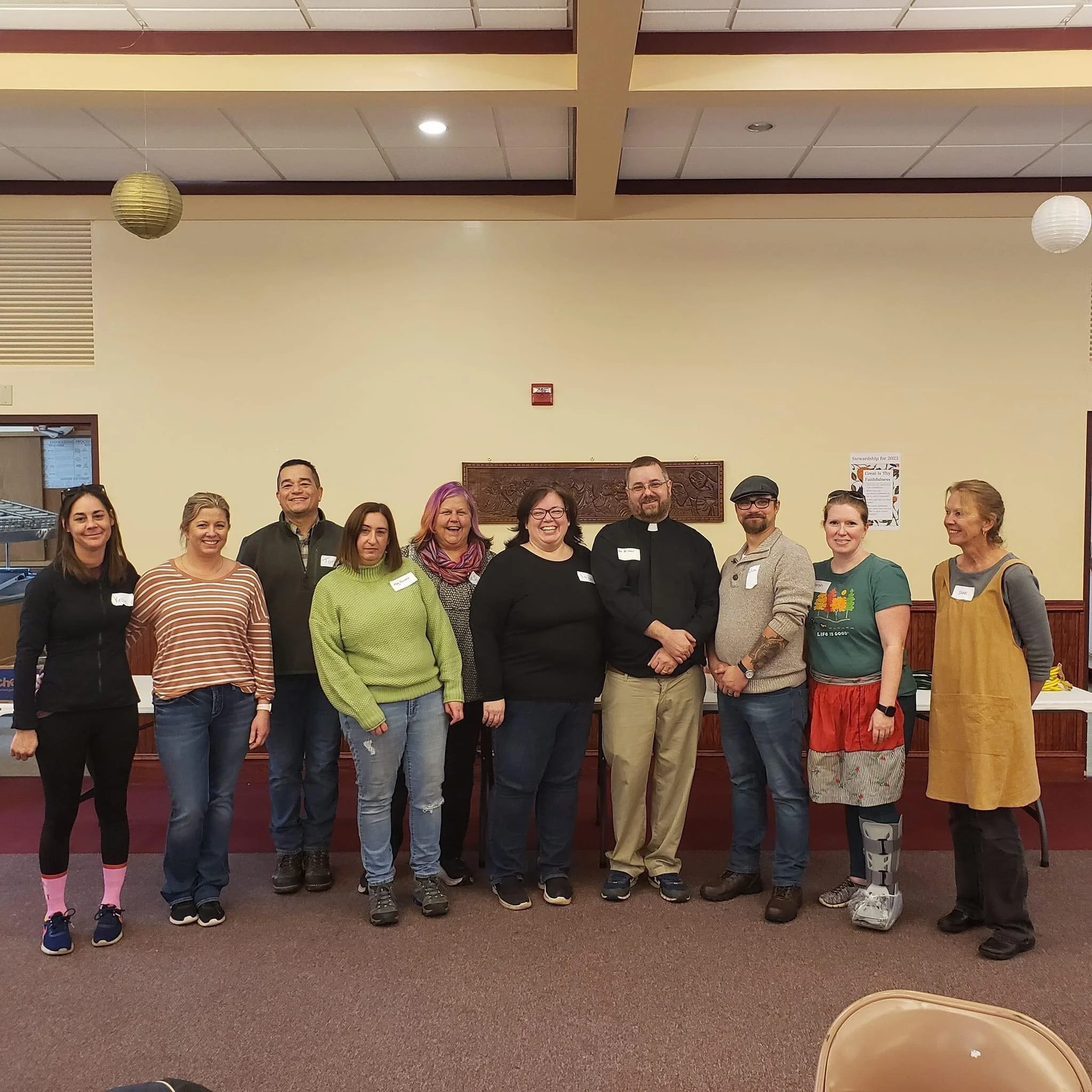 Group of 11 diverse people standing in a line indoors, smiling for a photo, with beige walls, wooden trim, and paper lanterns hanging from the ceiling.