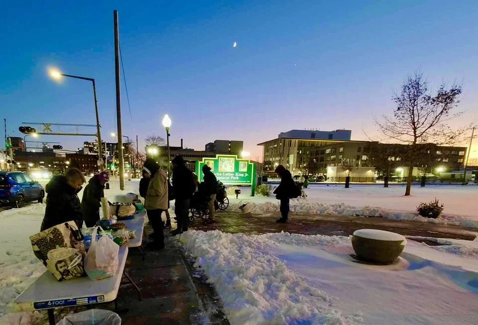 People gathering at an outdoor market in a snowy park during dusk, with some tables of items for sale, city buildings in the background, and a green illuminated sign.