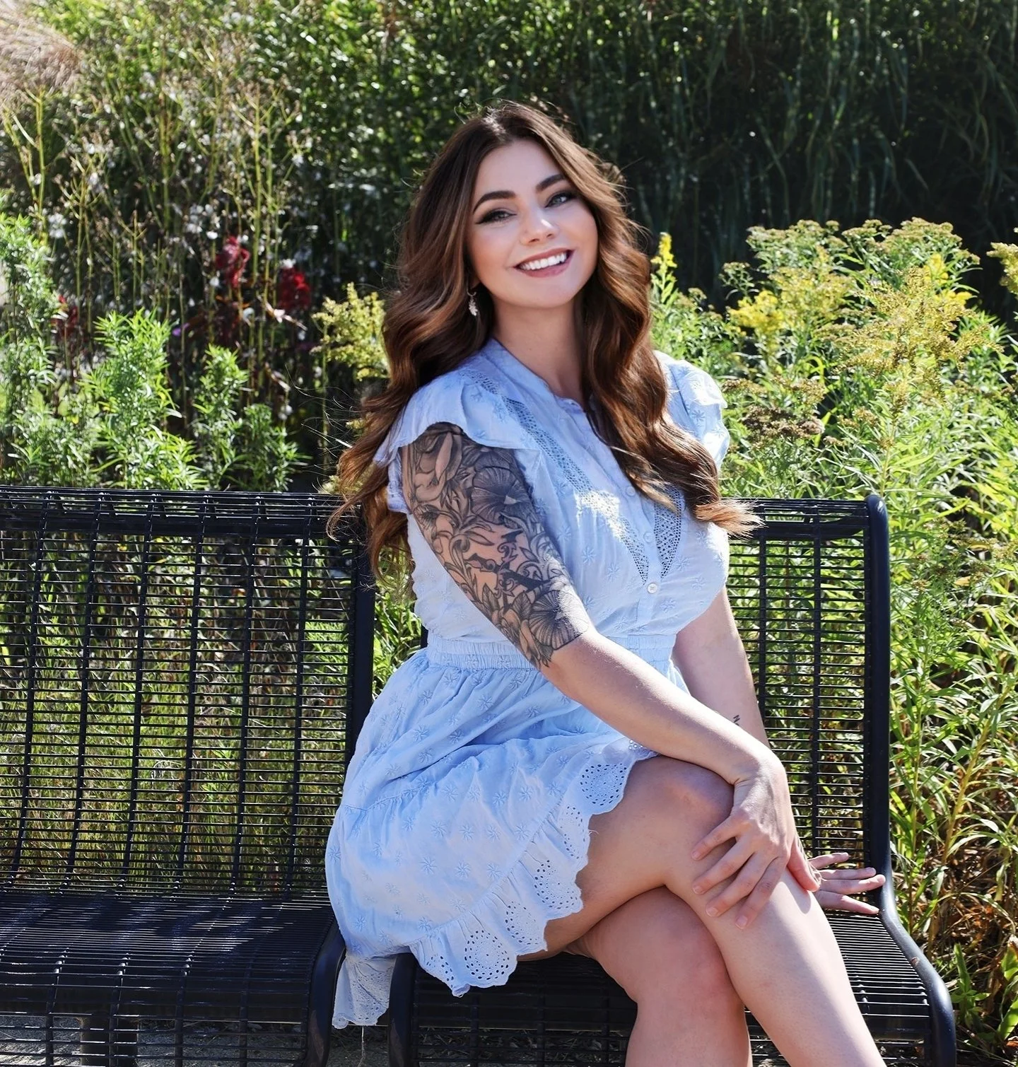 Young woman with long wavy brown hair, wearing a light blue dress with eyelet details, sitting on a black metal bench outdoors surrounded by green foliage, smiling at the camera.