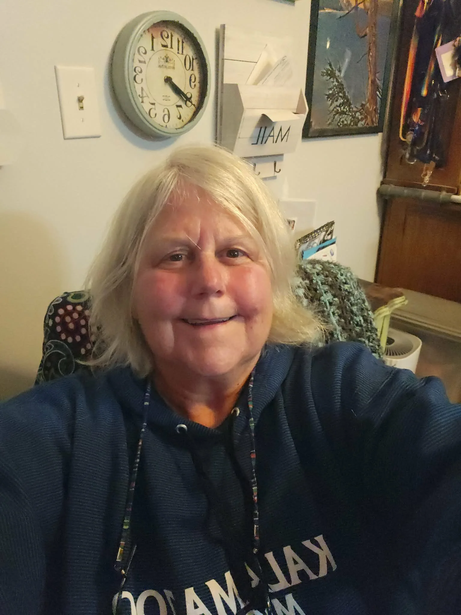 A smiling woman with shoulder-length blond hair taking a selfie indoors. Behind her is a white wall with a round clock, a light switch, and a wall-mounted box containing letters spelling 'MAIL'. There are also framed pictures and various items on shelves.