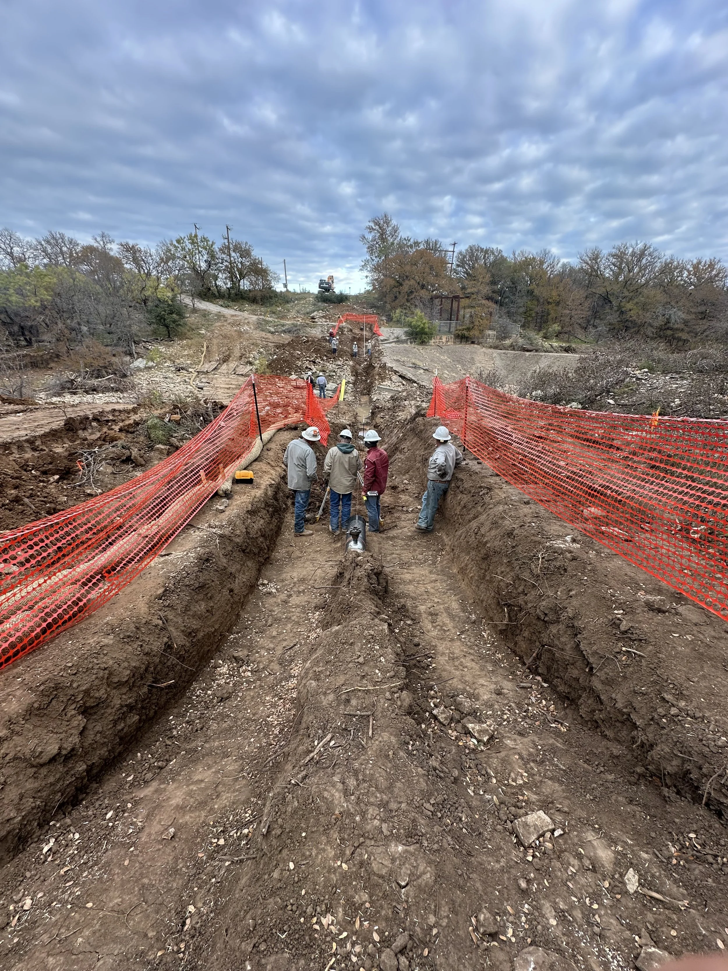 Construction workers in hard hats inspecting a deep excavation site with orange safety fencing, dirt trenches, and cloudy sky.