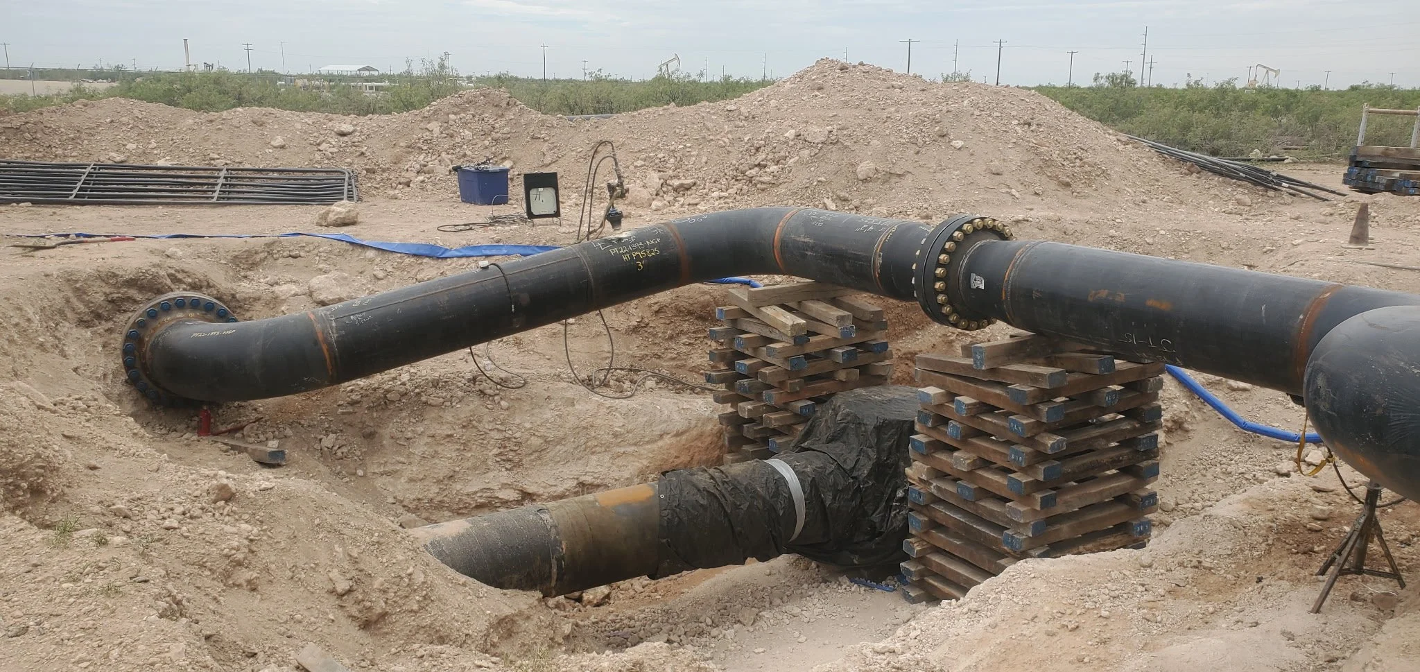 Underground pipeline construction site with large black pipes supported by wooden scaffolding and surrounded by dirt and gravel.