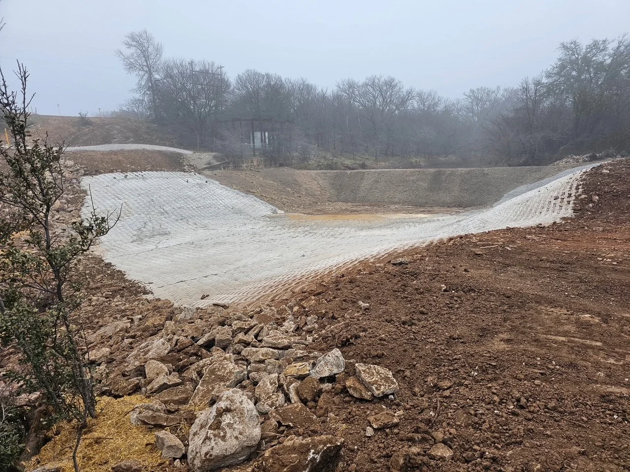 Construction site with a dirt slope, rocks, and a white geogrid liner possibly for erosion control, with leafless trees and overcast sky in the background.