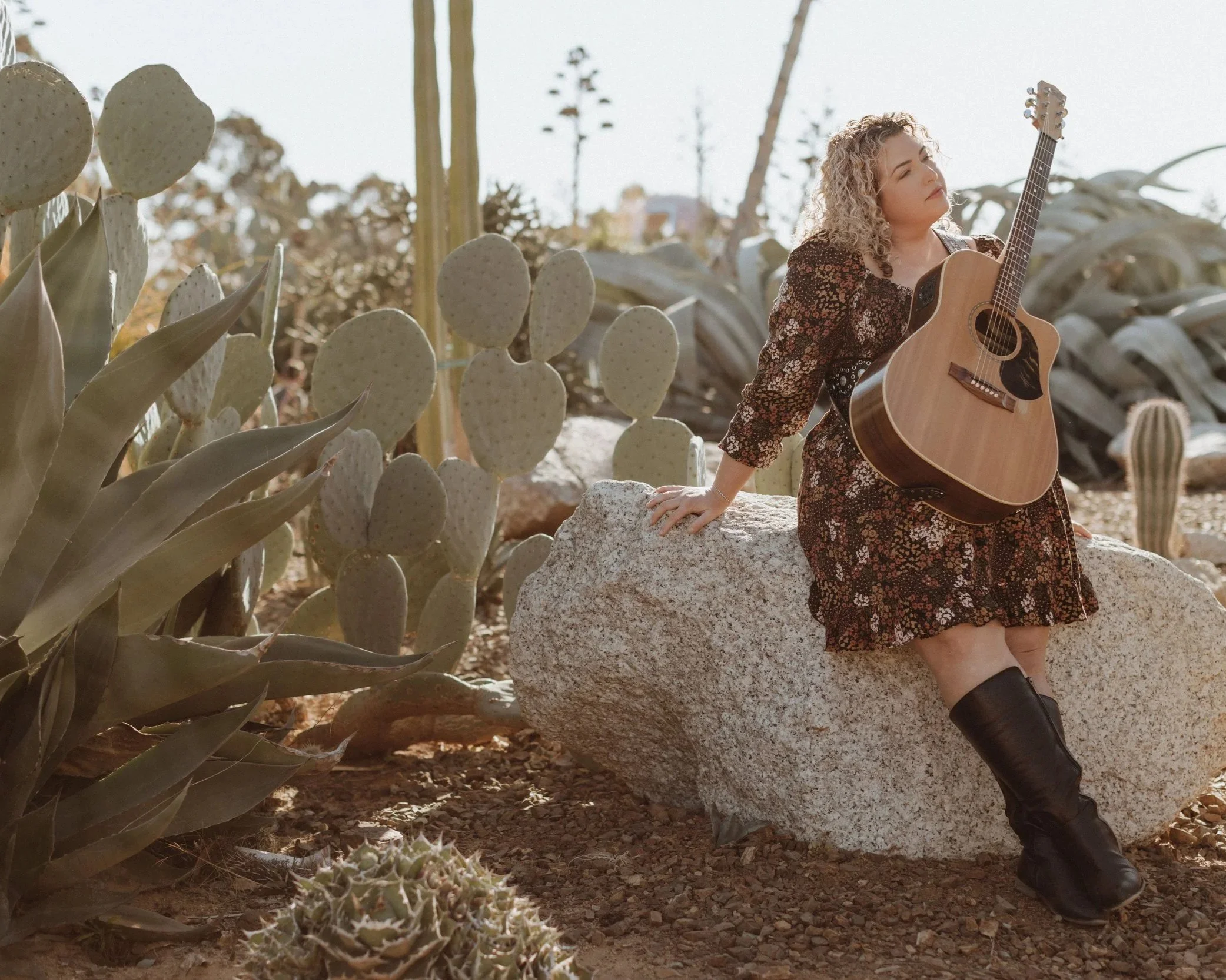 A woman with curly blonde hair sitting on a large rock in a desert, holding an acoustic guitar, surrounded by cacti and succulents.