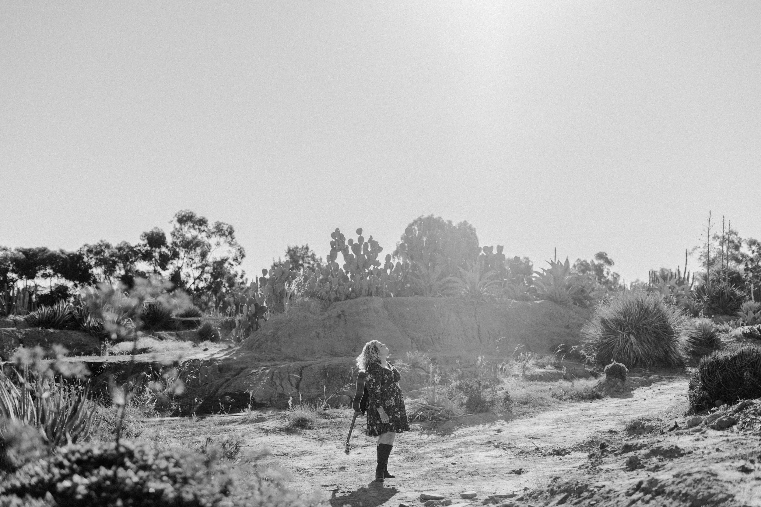 A woman in a floral dress and tall boots walking through a desert landscape with cacti and succulents, holding a guitar on her back, in black and white.
