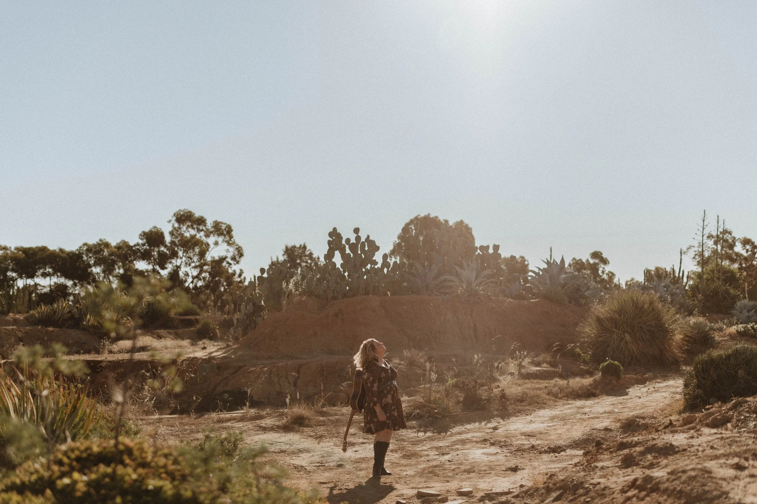 A woman walking on a dirt trail in a desert landscape with cacti and sparse vegetation, under a bright sky.