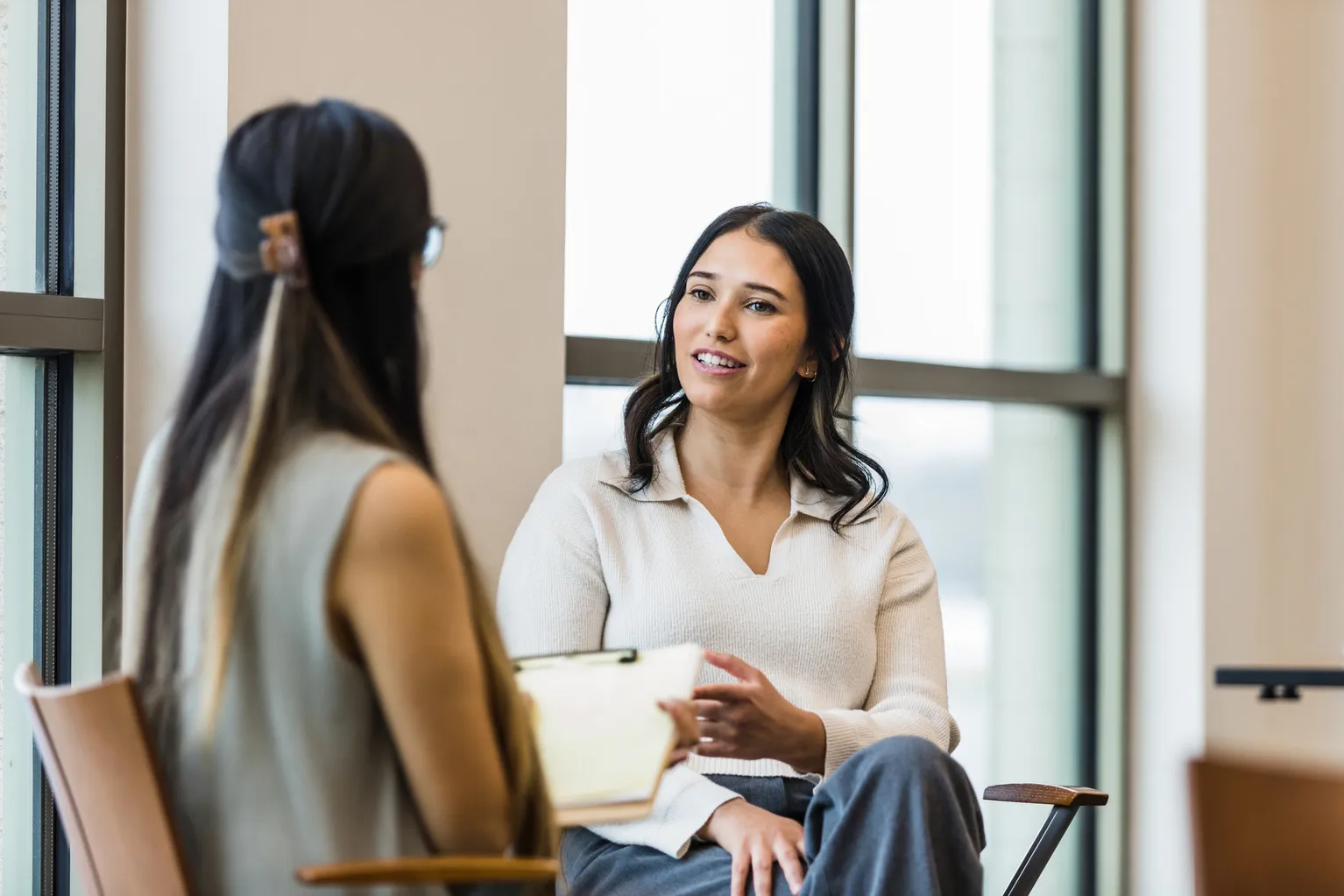 Two women having a conversation in a room with large windows, one woman is holding a notebook.