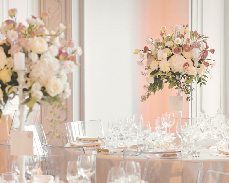 Elegant event tables set with glassware, napkins, and floral centerpieces in a bright room with large windows.