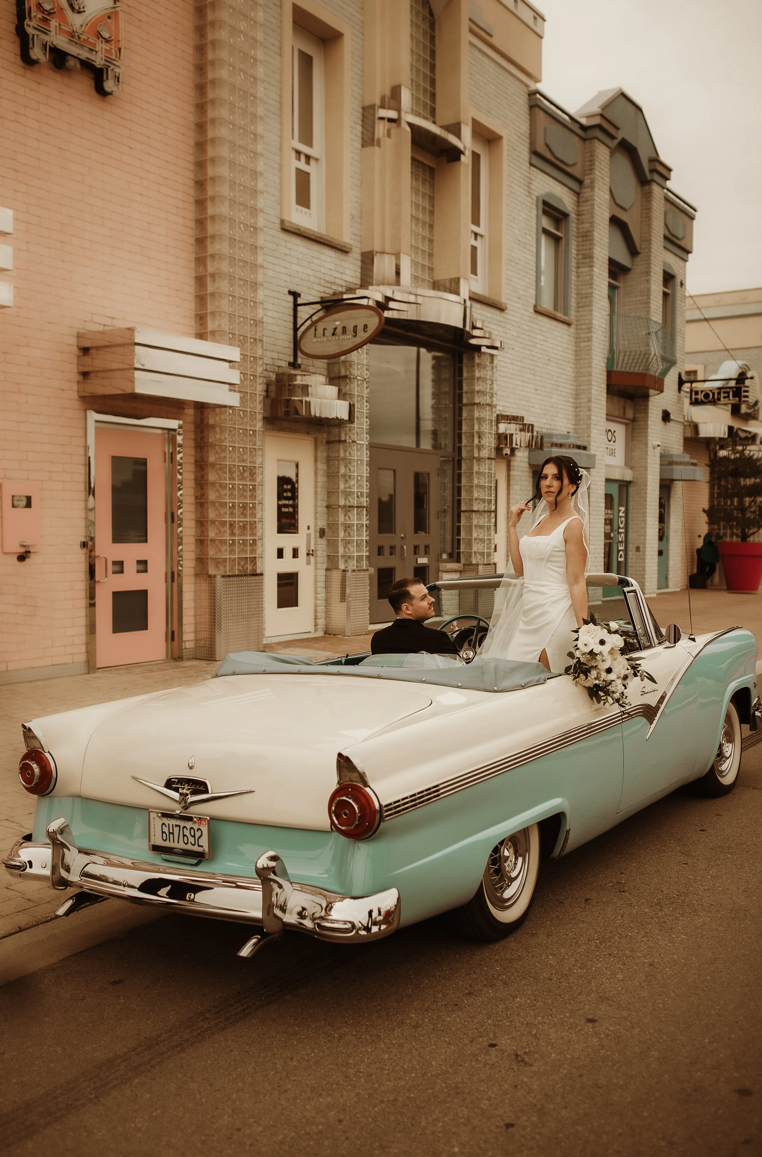 A bride in a white wedding dress standing in a vintage convertible car with a groom sitting in the driver's seat, on a city street with buildings and signs in the background.