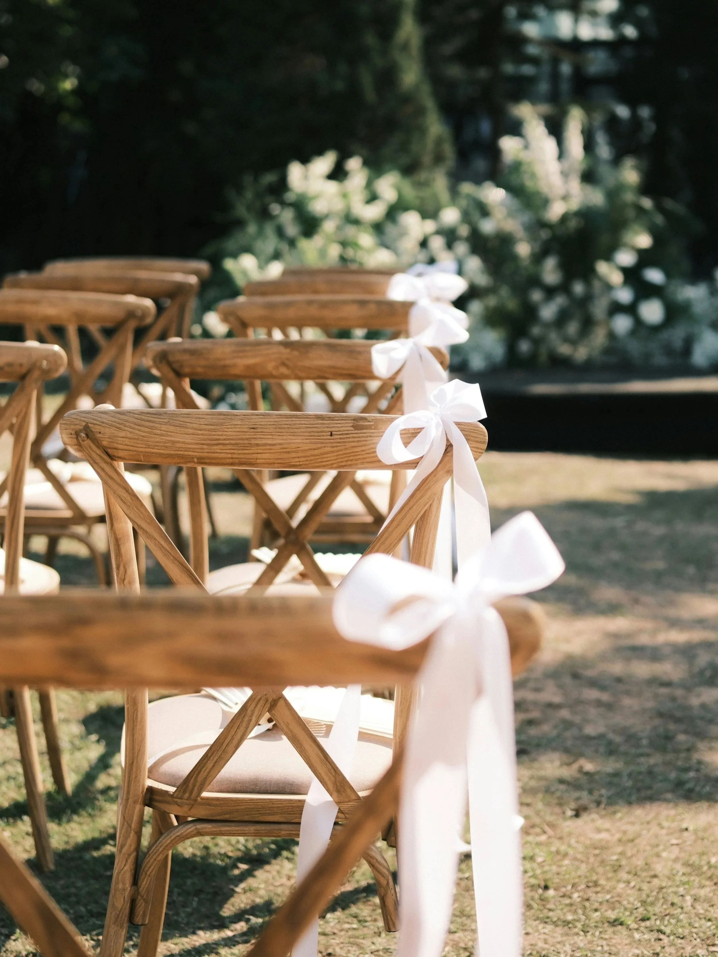 A row of wooden chairs decorated with white ribbons, set outdoors, with greenery in the background.