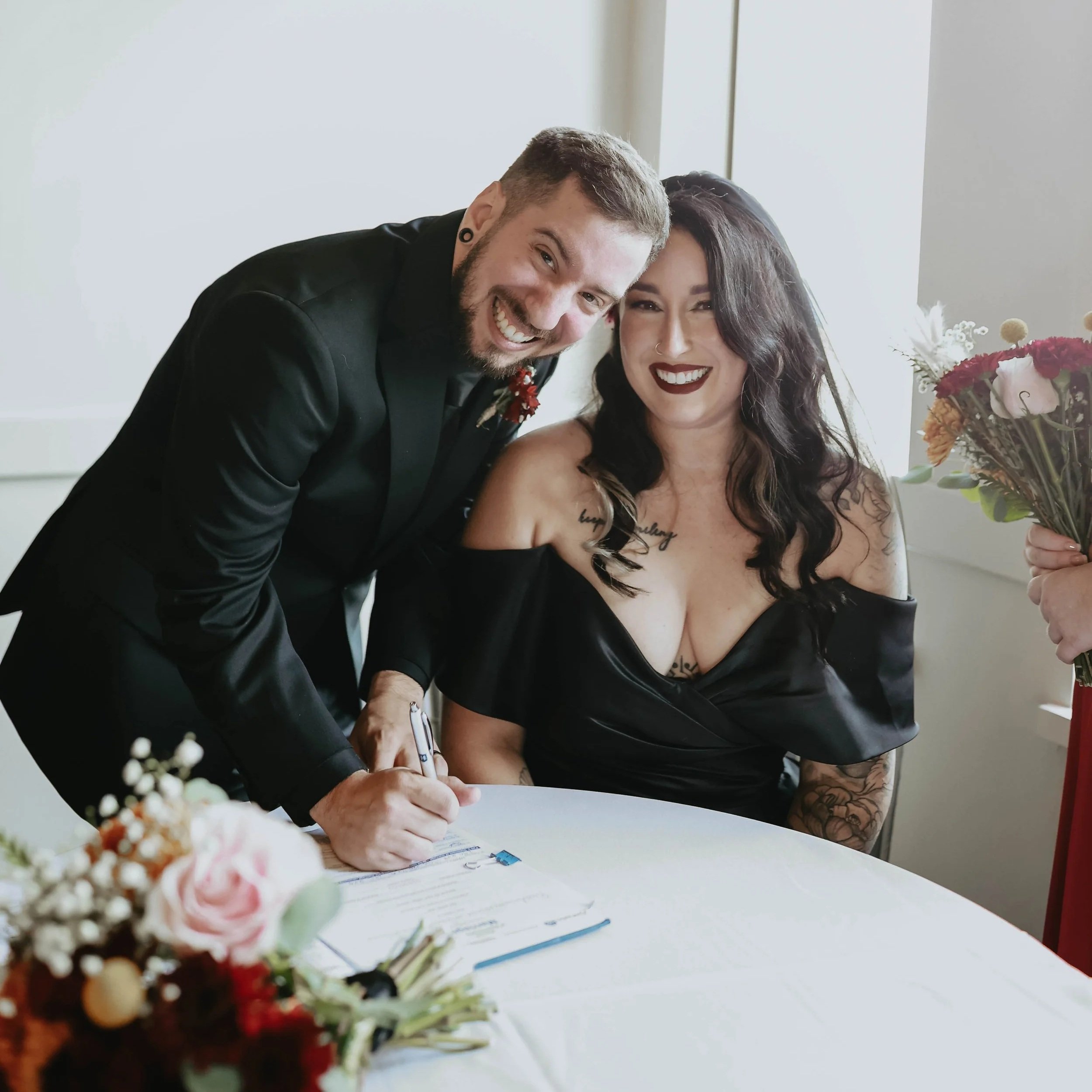 A man and woman at a wedding, signing a document at a table. The woman is holding a bouquet of flowers, and the man is signing a paper with a pen.
