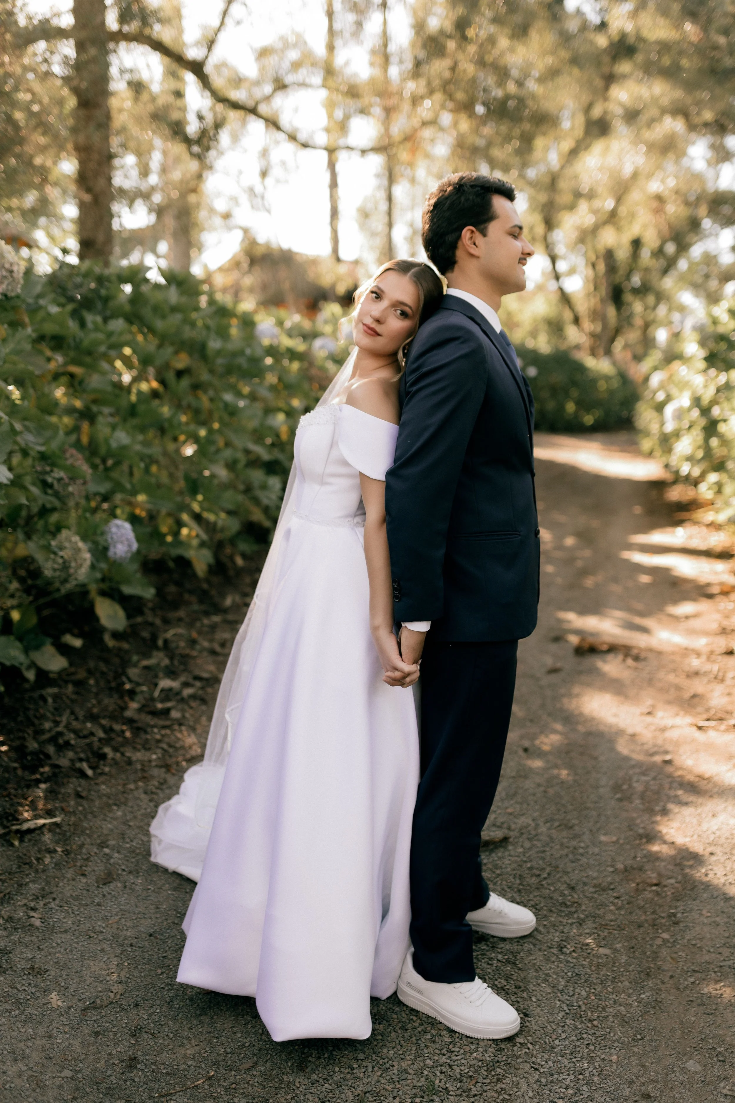 A bride and groom stand back to back outdoors, holding hands, with the bride resting her head on the groom's shoulder. The bride wears a white off-the-shoulder wedding gown and the groom is dressed in a navy blue suit. They are surrounded by greenery and trees with sunlight filtering through.