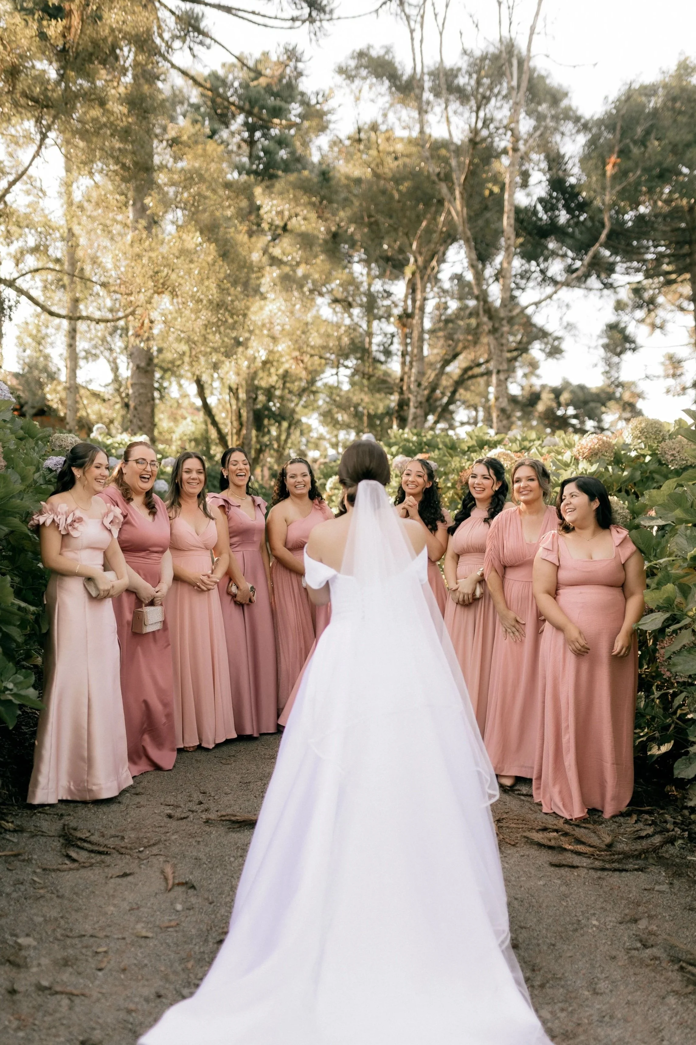 A bride in a white wedding gown standing outside in front of nine bridesmaids dressed in pink dresses, surrounded by greenery and tall trees, smiling and laughing.