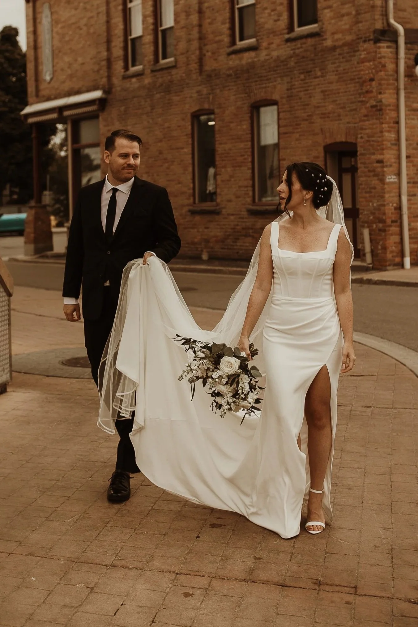 A bride and groom walking on a city sidewalk during their wedding. The groom is dressed in a black tuxedo, and the bride is wearing a white wedding gown with a slit up the leg, carrying a bouquet of flowers, and wearing white open-toe heels. The background features a brick building with multiple windows.