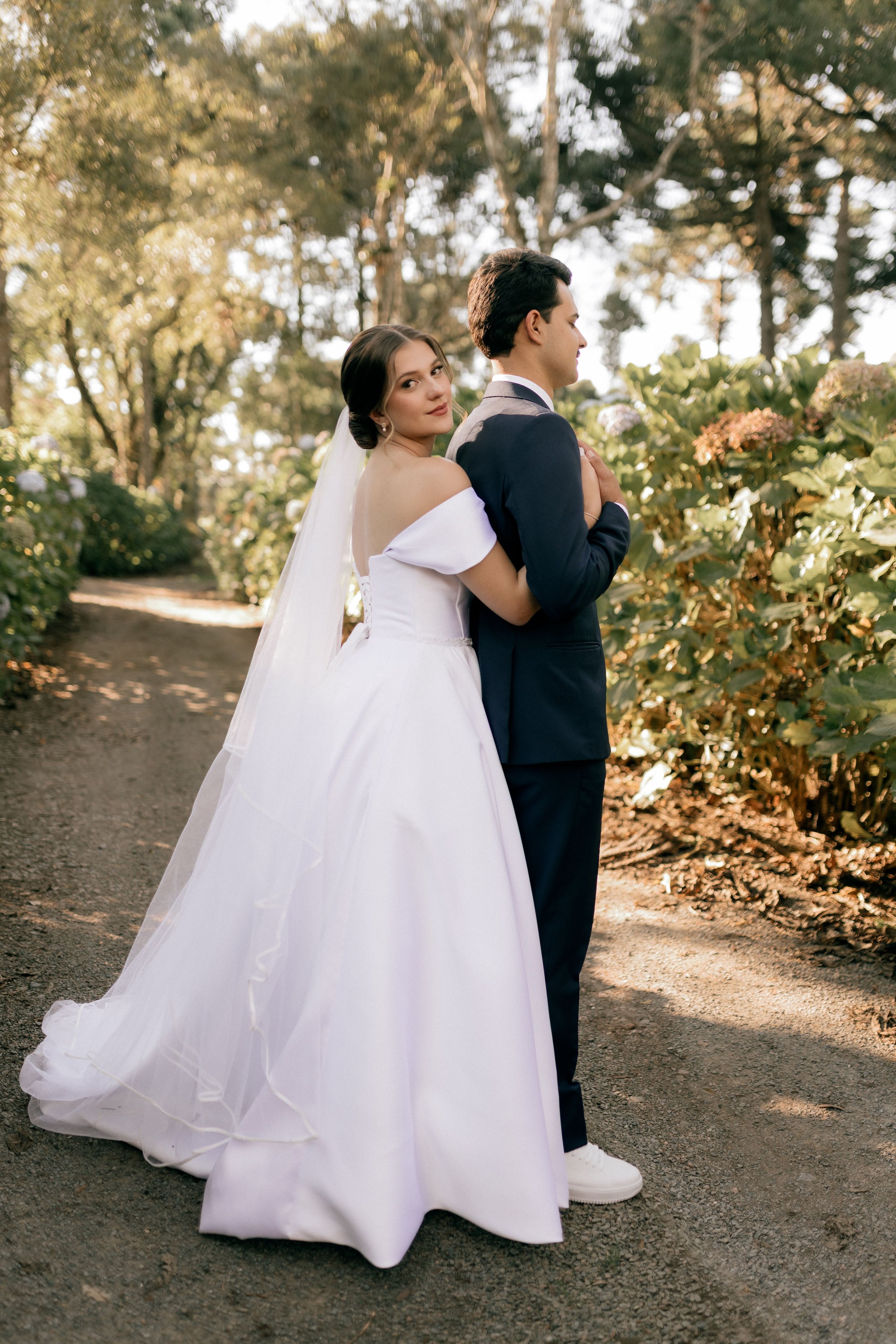 A bride and groom standing back-to-back outdoors in a wooded area during daylight. The bride wears a strapless white wedding gown with a long veil, and the groom wears a dark suit with a white shirt. The bride looks over her shoulder with a slight smile, and the groom looks away with a serious expression.