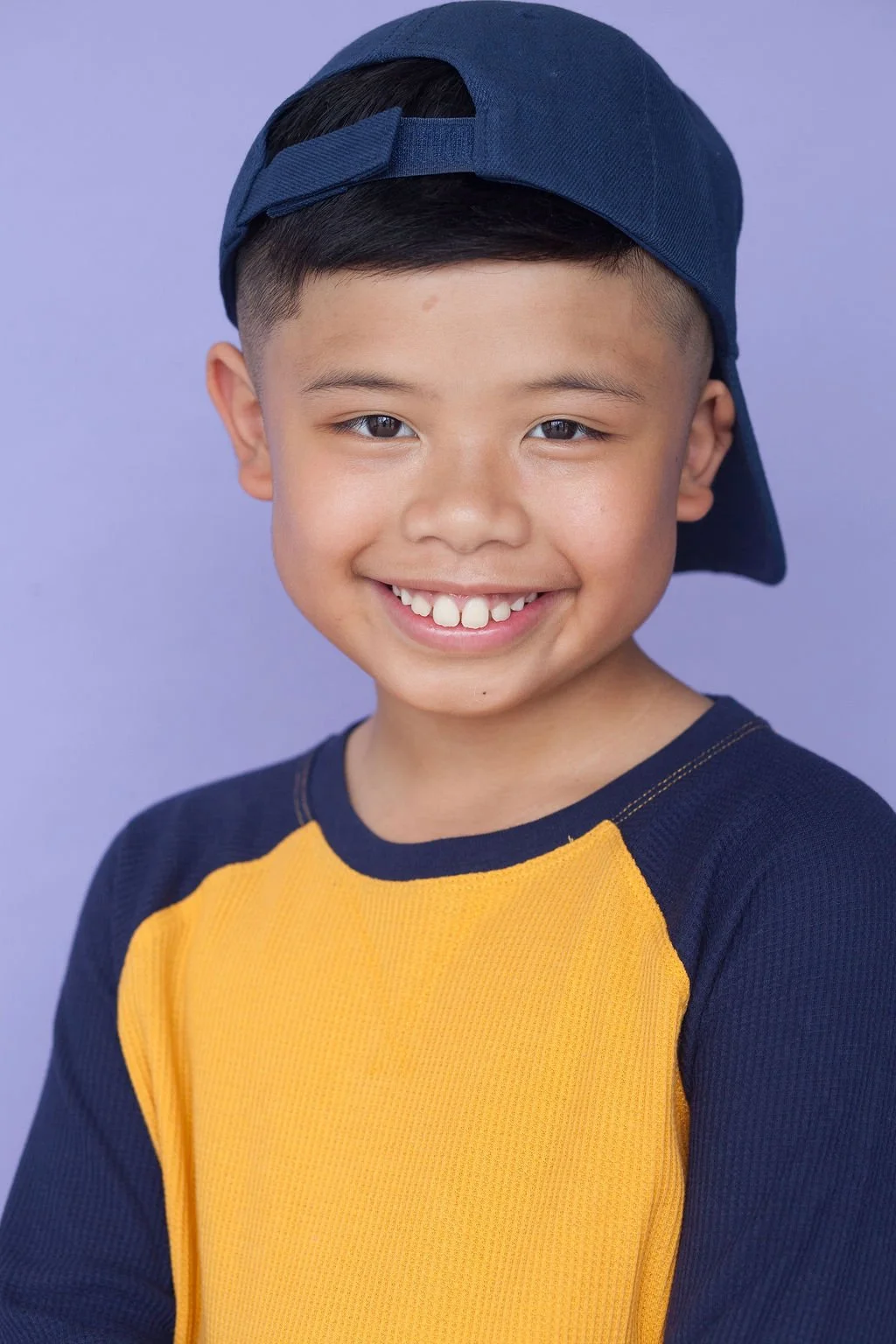 A smiling young child actor posing in front of a light blue backdrop wearing a baseball cap backwards and a long sleeve yellow and blue top.