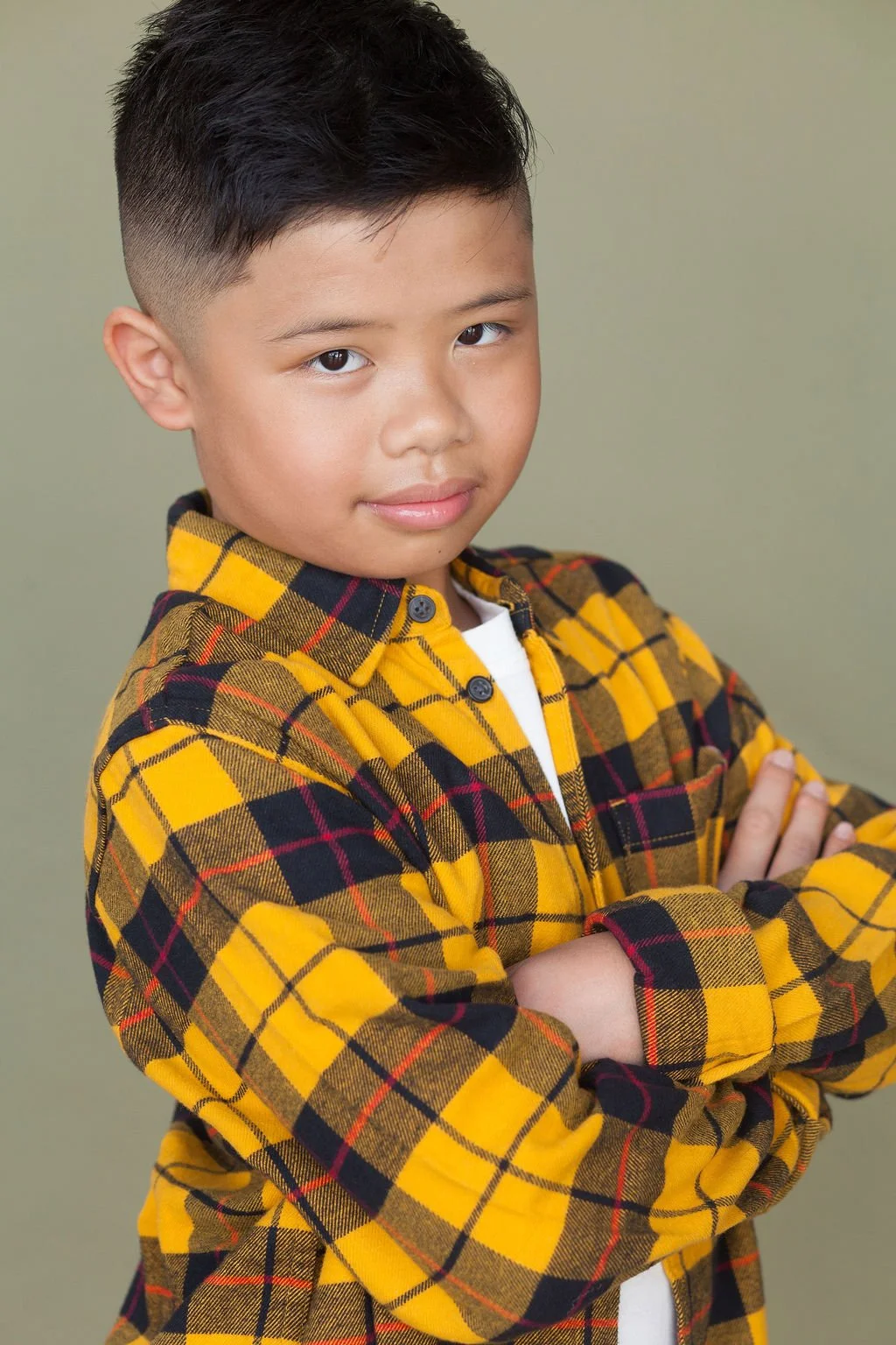 A child actor standing on their side profile with their arms folded across their chest wearing a yellow collared flannel in front of neutral olive colored backdrop.