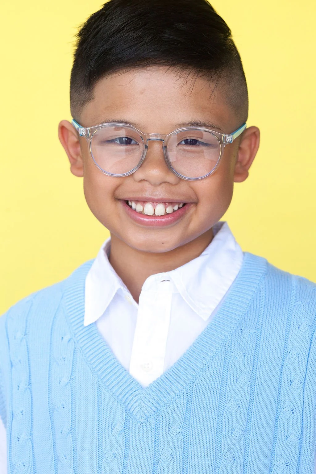 Photo of a child actor standing in front of yellow backdrop wearing eyeglasses and a light blue sweater vest over a white button-collared top.