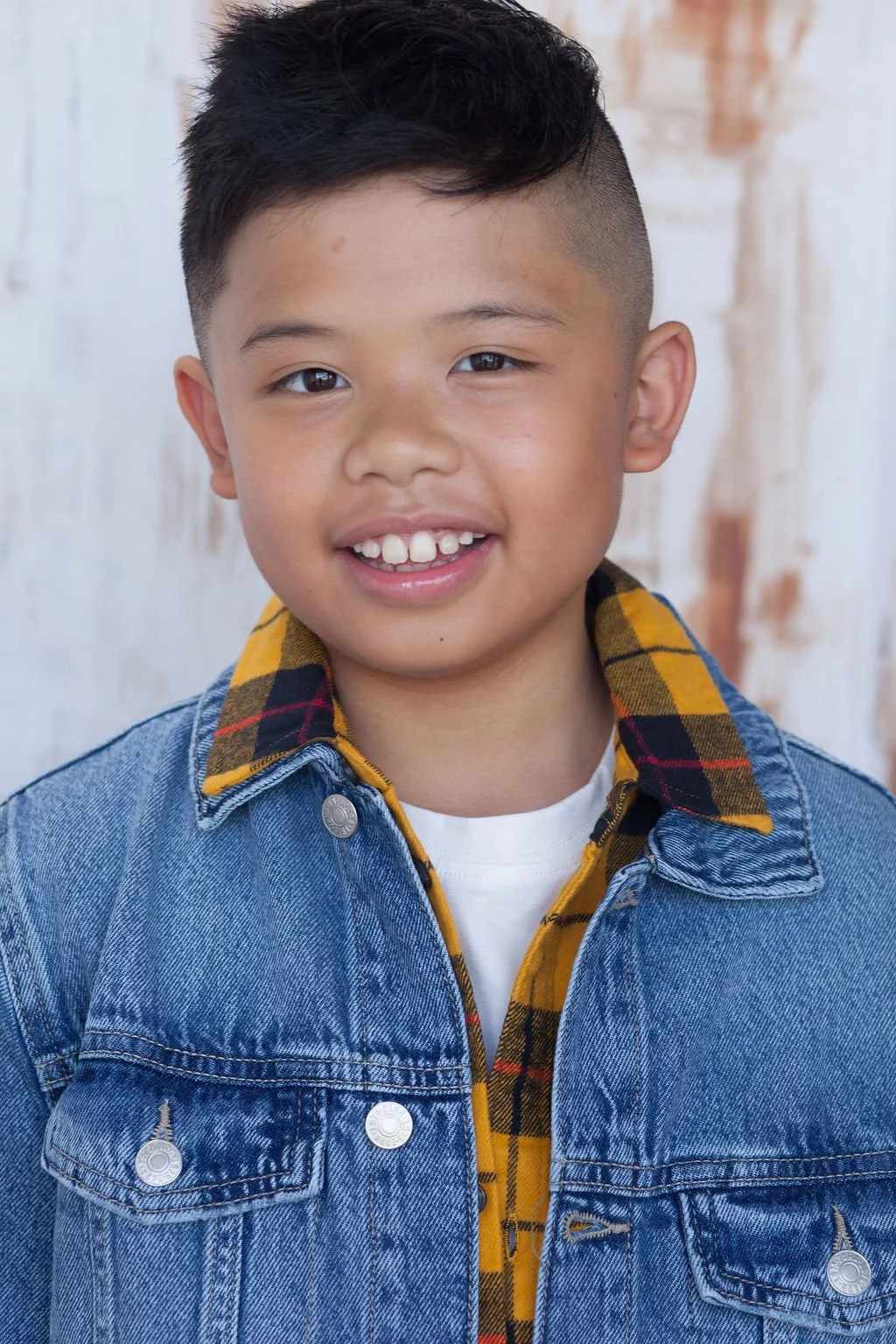 A child actor with a blue denim jacket and a yellow collared flannel shirt with a white t-shirt underneath posing in front of an industrial backdrop.