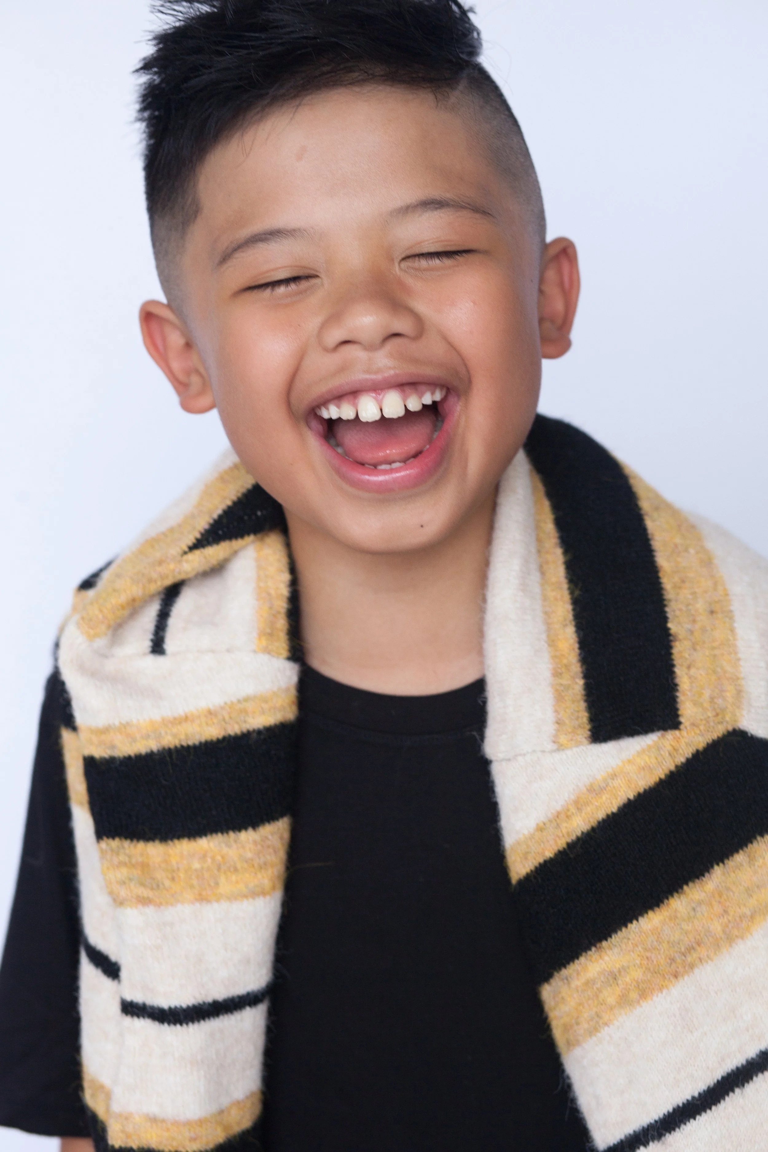headshot of a boy laughing with eyes closed wearing a black top with a sweater wrapped around the neck behind a off-white backdrop.
