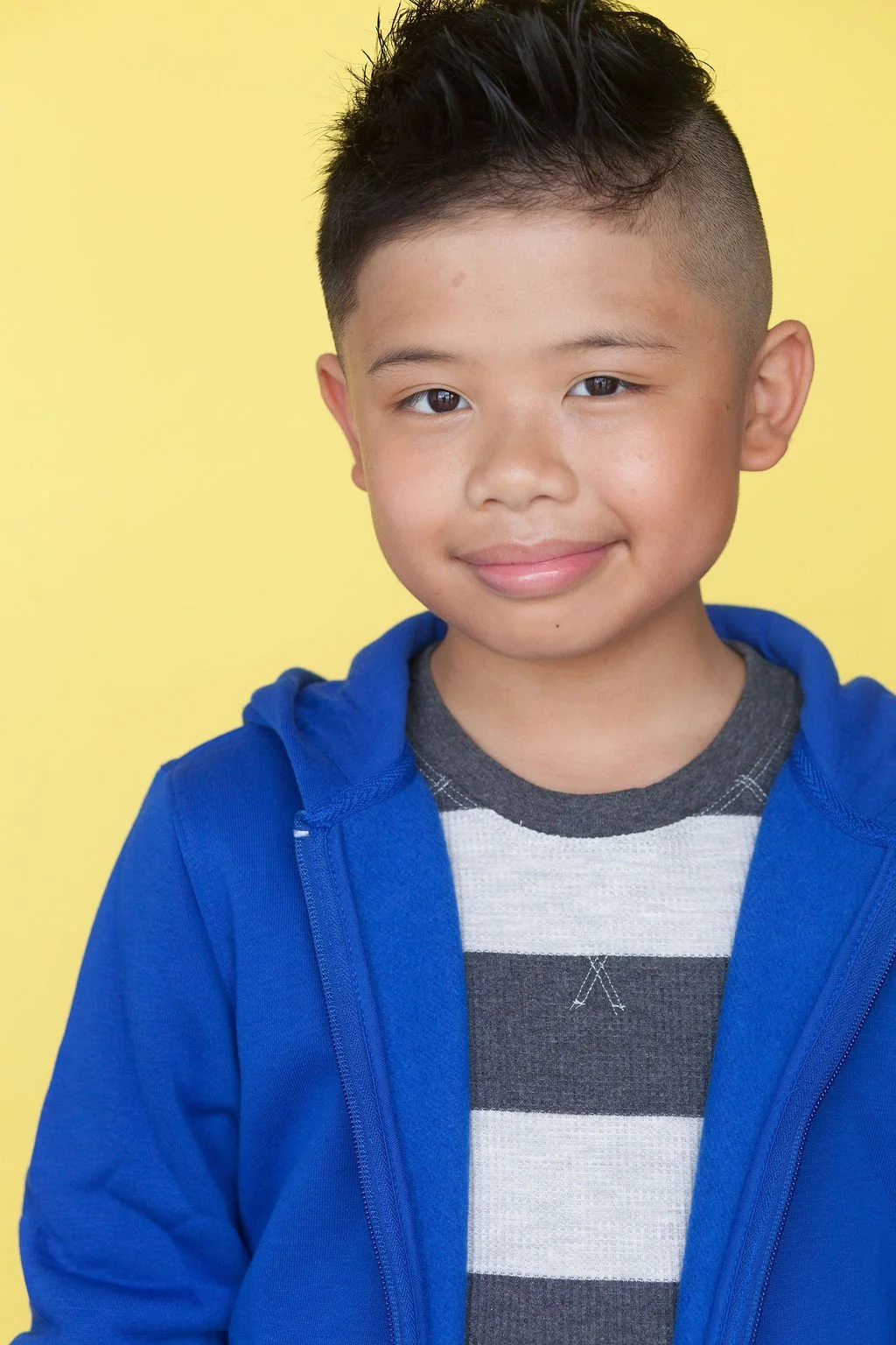 A child actor smiling in front of a yellow backdrop wearing a blue hoodie with a white and grey striped long sleeve top underneath.
