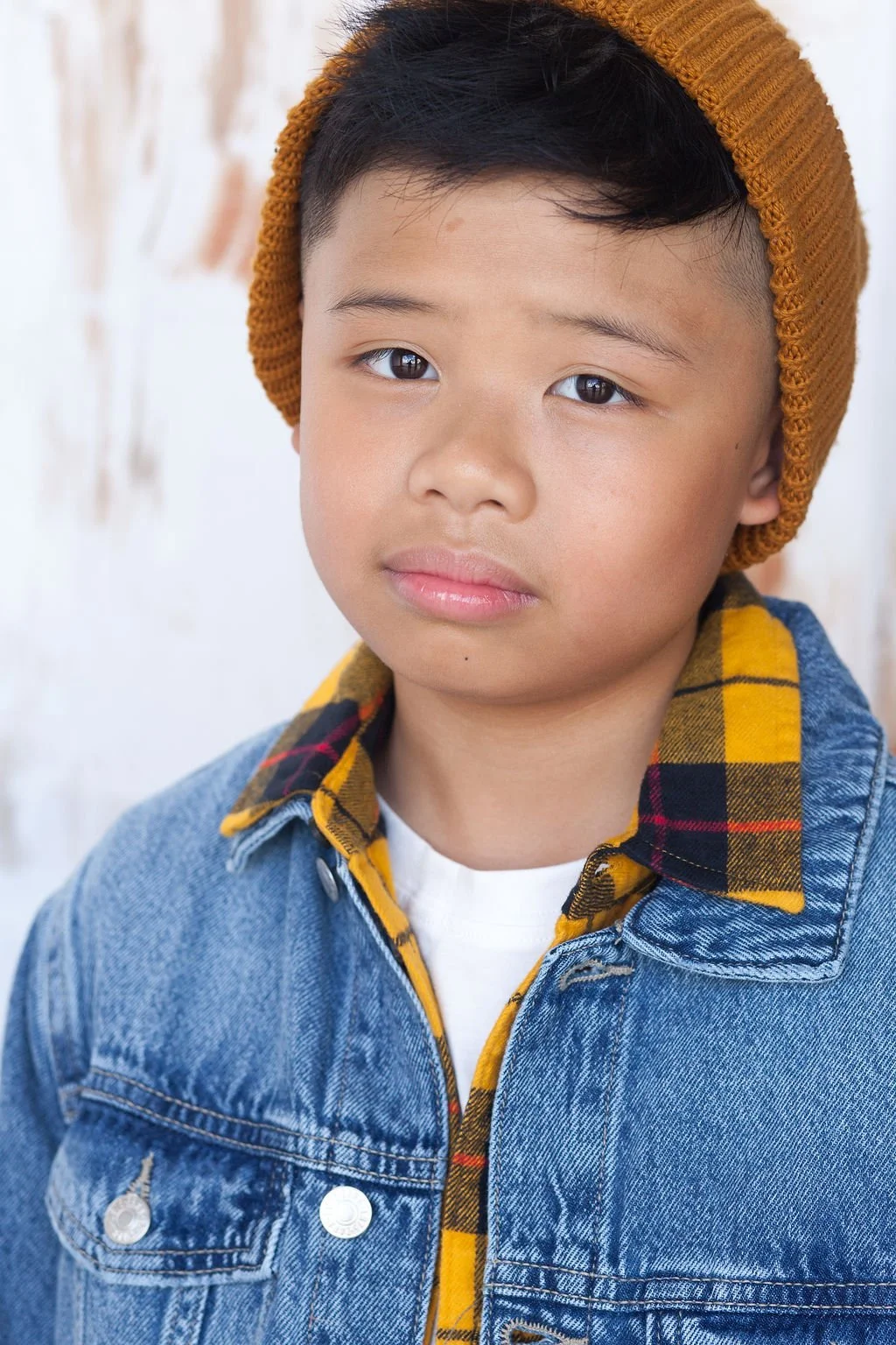 Child actor facing forward in front of an industrial wall wearing a brown beanie hat and a denim jacket with a yellow flannel collared shirt and a white t-shirt underneath.