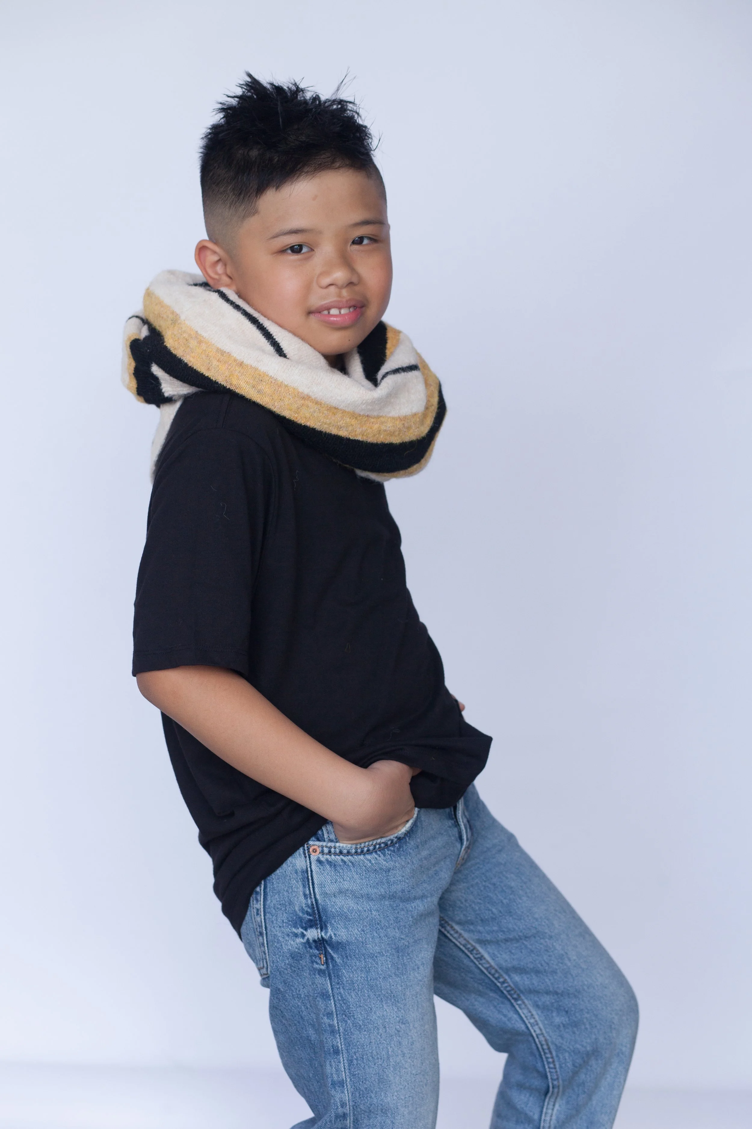 A young actor profile posing with their hands in their pockets wearing a black t-shirt and blue denim jeans with a sweater wrapped around their neck.