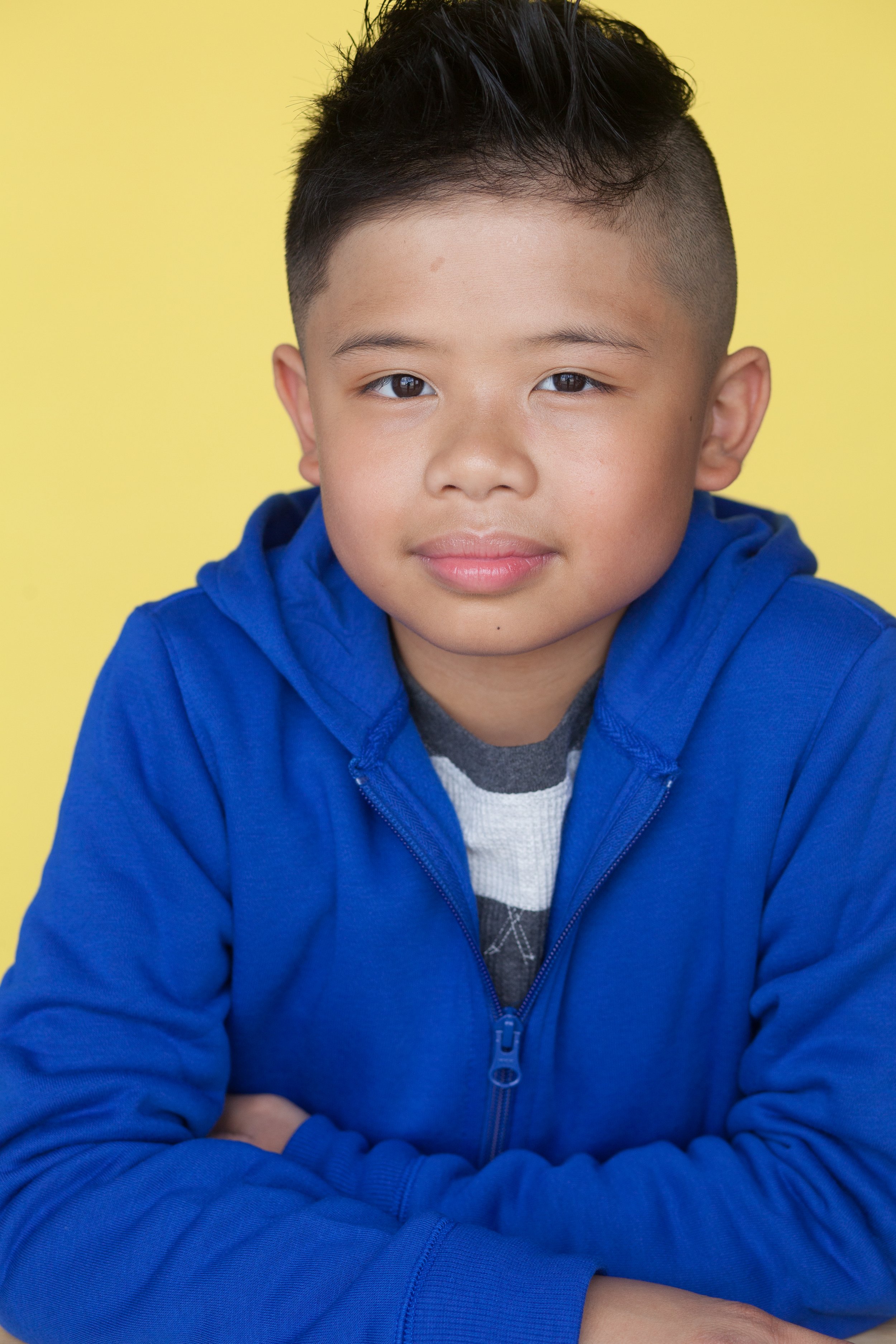 A child actor pacing forward with their arms folded across their chest on a table posing in front of a yellow backdrop  wearing a blue hoodie jacket and a white and gray striped top.