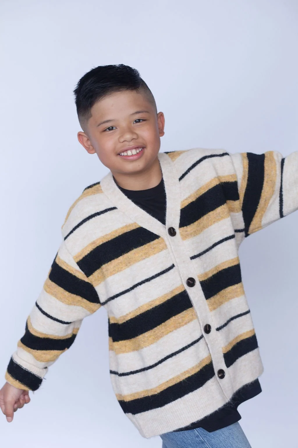 A young kid smiling, posing and standing with their arms wide open in front of a light grey backdrop wearing a long sleeved striped light beige button-up sweater with a black t-shirt underneath.