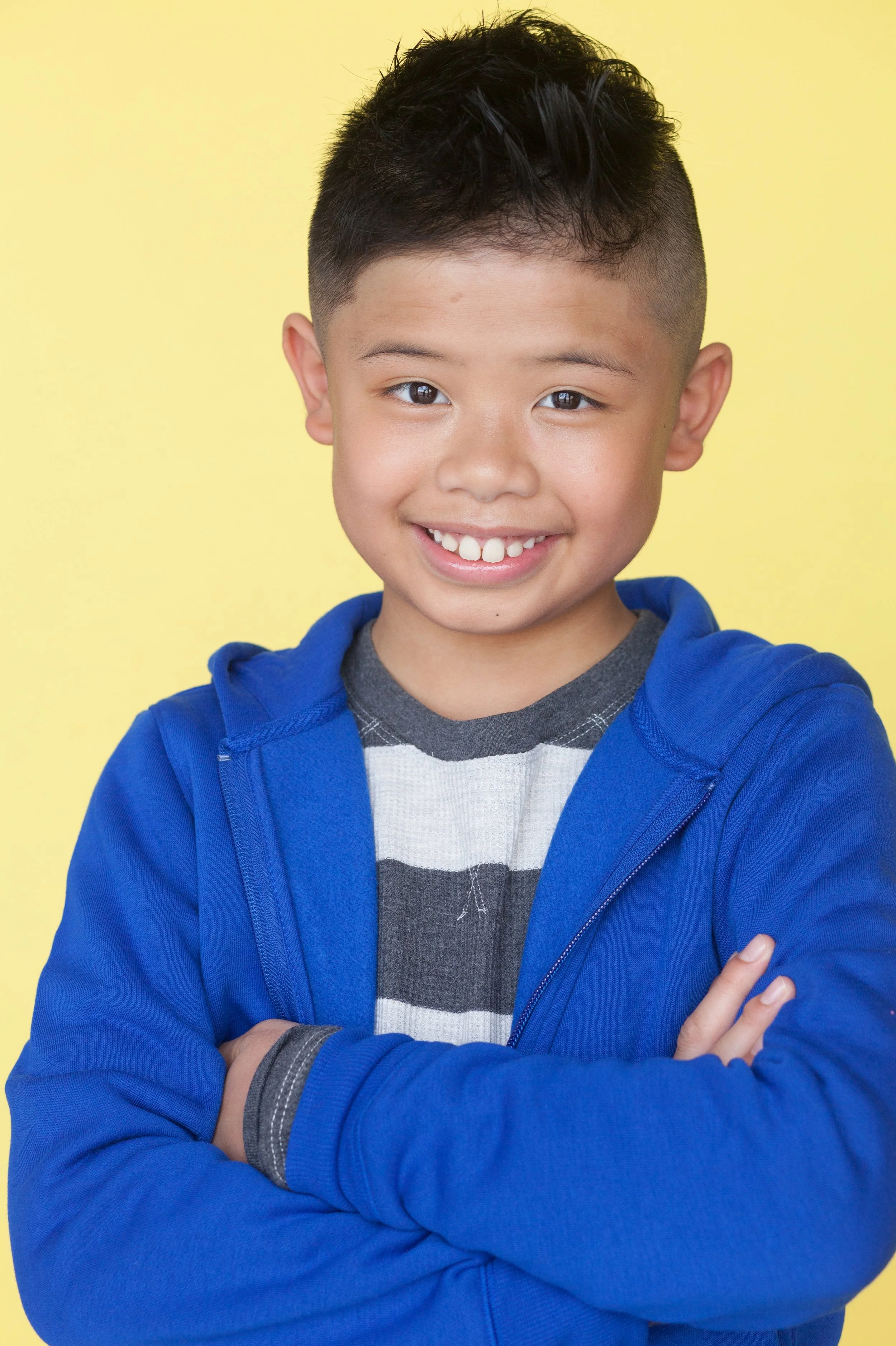 Photo of child actor smiling and facing forward behind a yellow backdrop wearing a blue hoodie jacket and a striped grey and white long sleeve shirt with their arms folded across their chest.