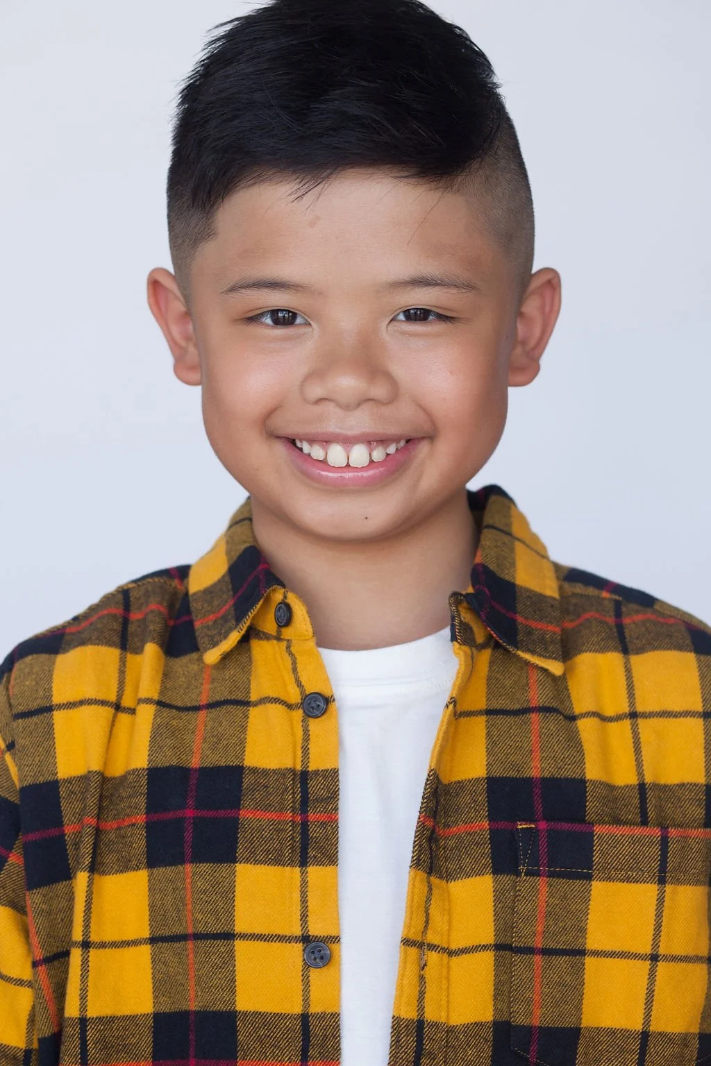 Child actor smiling facing forward in front of a light grey backdrop wearing a yellow collared flannel shirt and a white t-shirt underneath