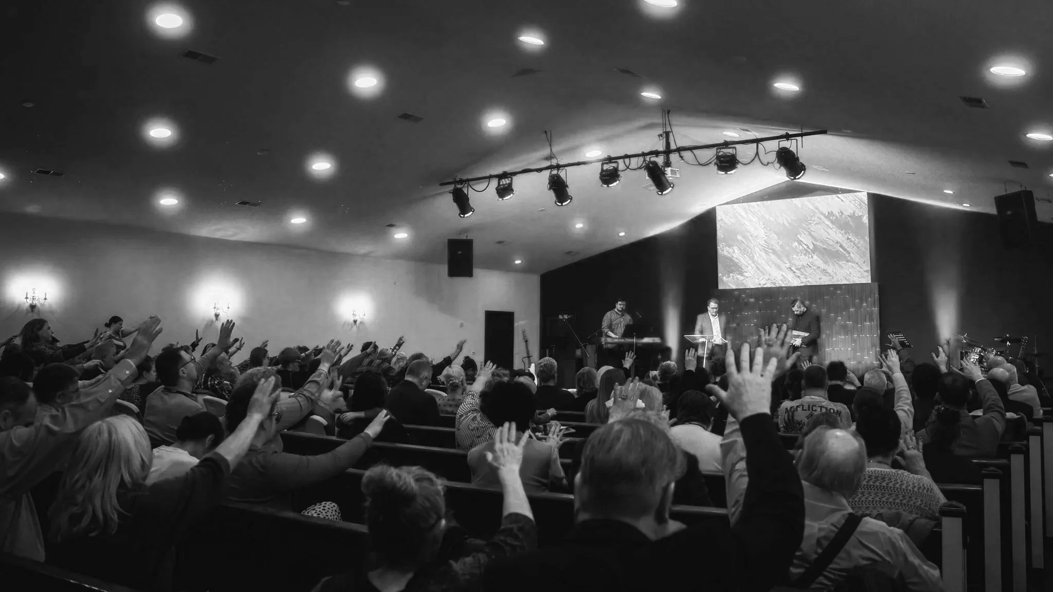 Black and white photo of a large congregation in a church or auditorium, with many people raising their hands while a band performs on stage with instruments and microphones.