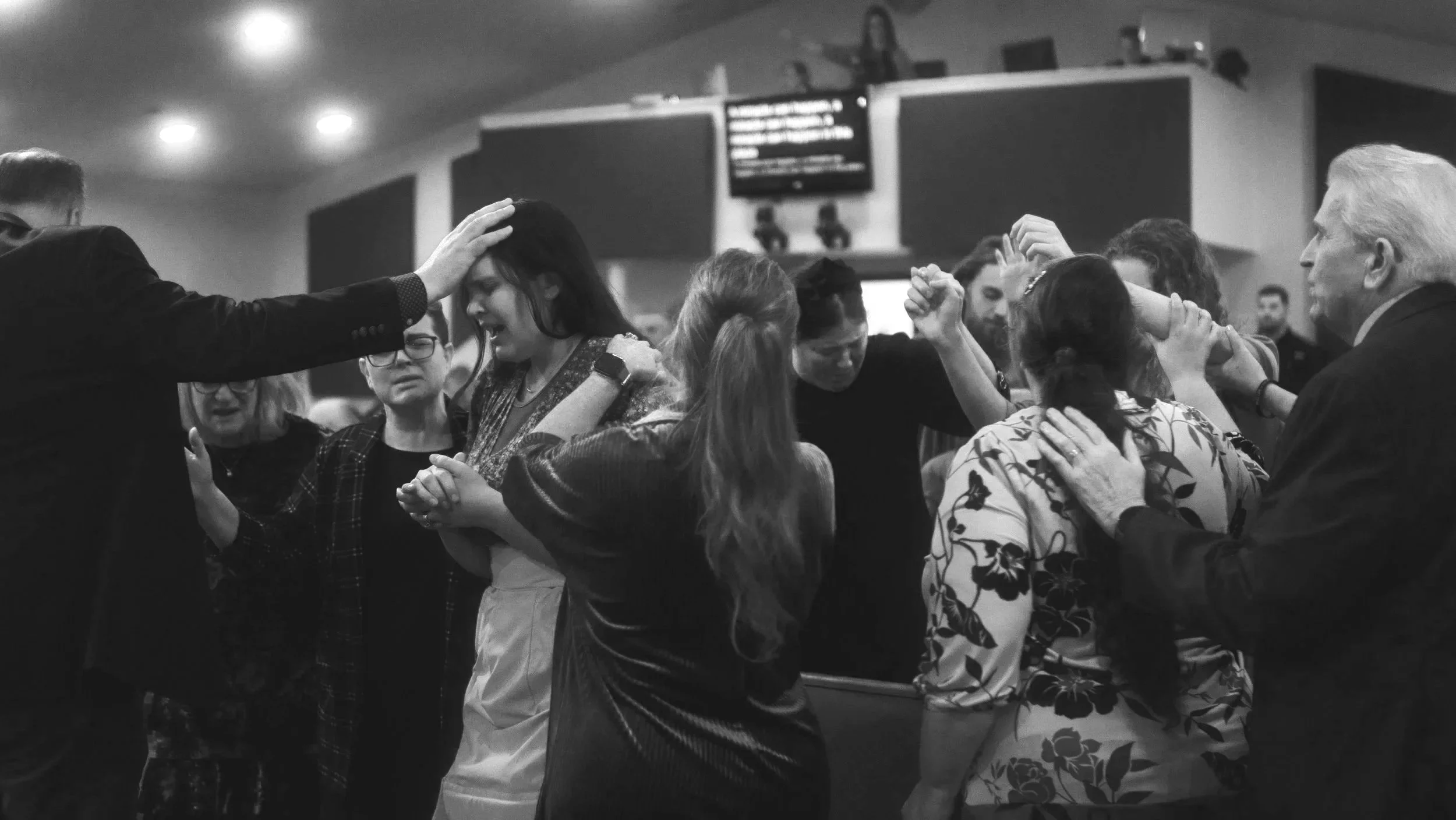 A black and white photo of a group of people in a church or religious gathering, with some holding hands and praying, while one person is being anointed or blessed by a man in a suit.