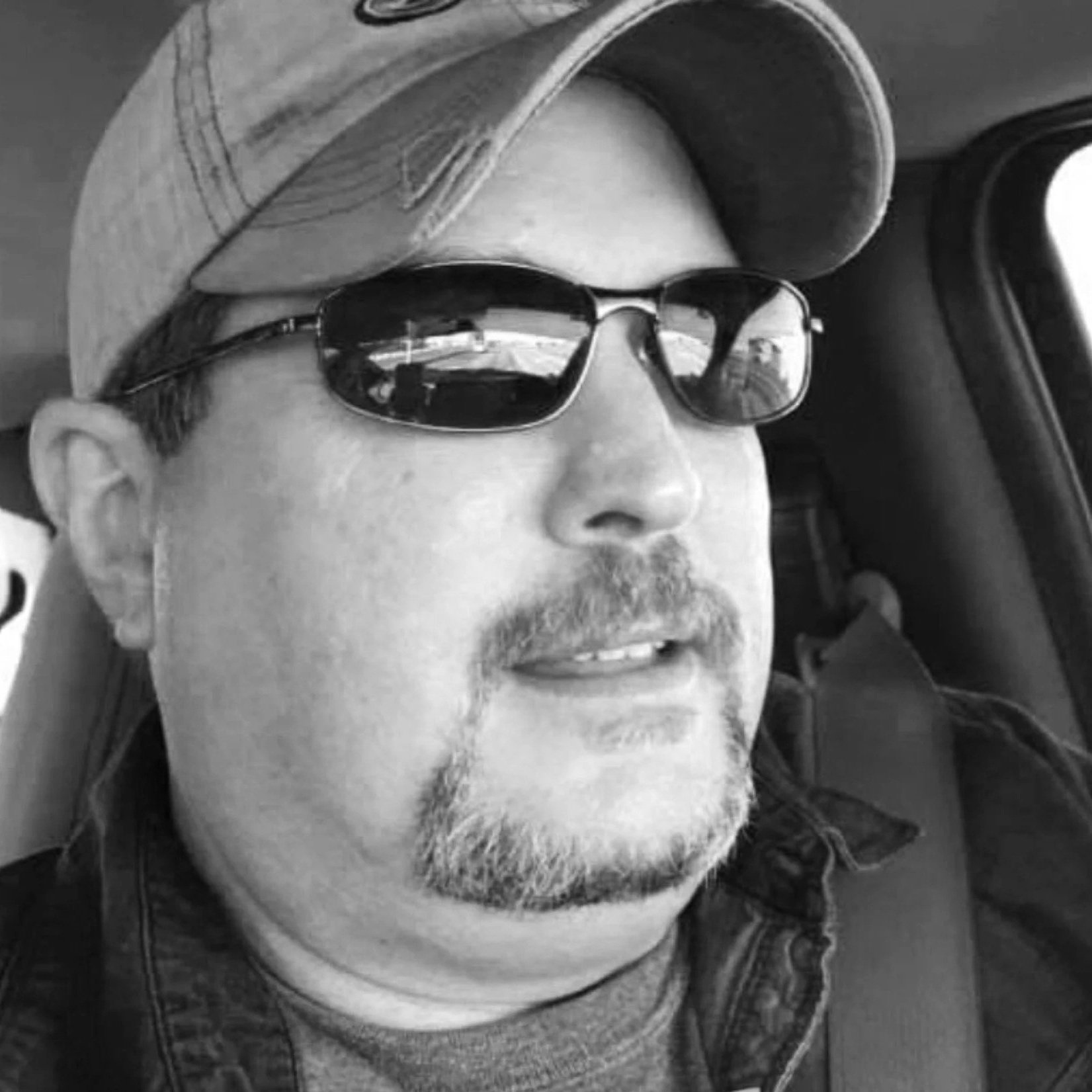 A man wearing sunglasses, a baseball cap, and a denim jacket sitting in a vehicle.