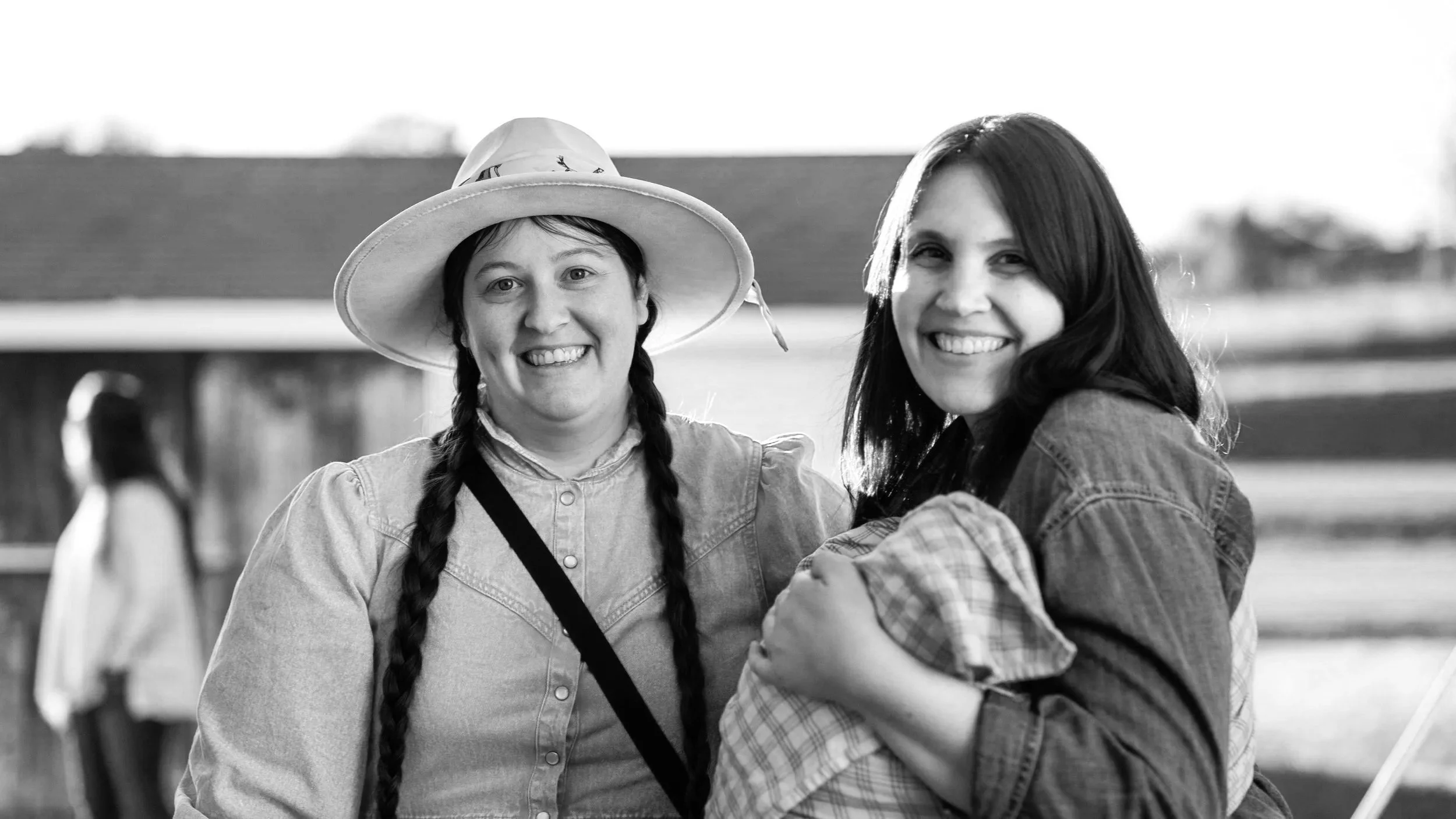 Two smiling women with braids, one with a wide-brimmed hat, stand outdoors, with a blurred person and a building in the background.