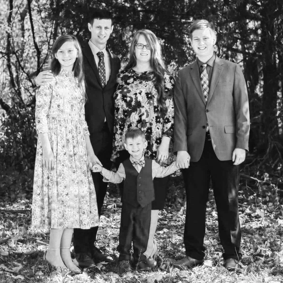 Family outdoors, group of six people, including four adults and two children, posing together, holding hands, surrounded by trees and fallen leaves, black and white photograph.