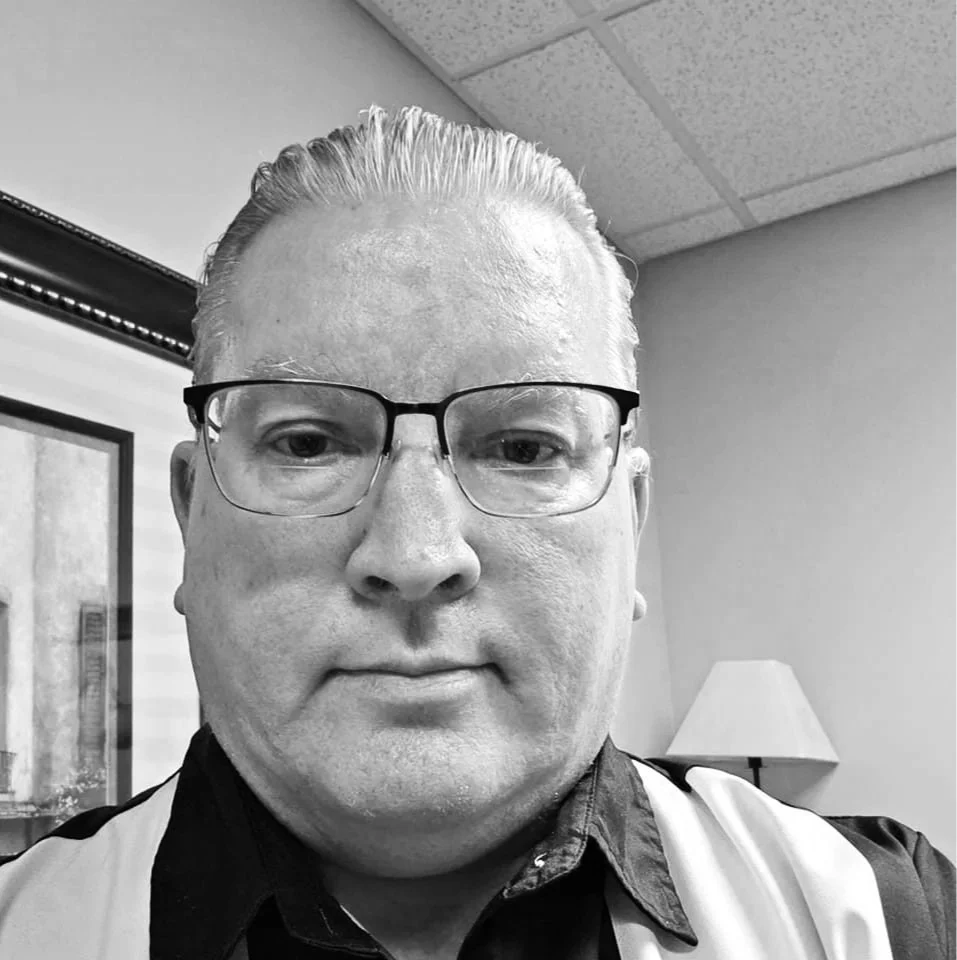 A man wearing glasses and a black shirt with a white vest, indoors with a lamp and picture frame visible in the background.