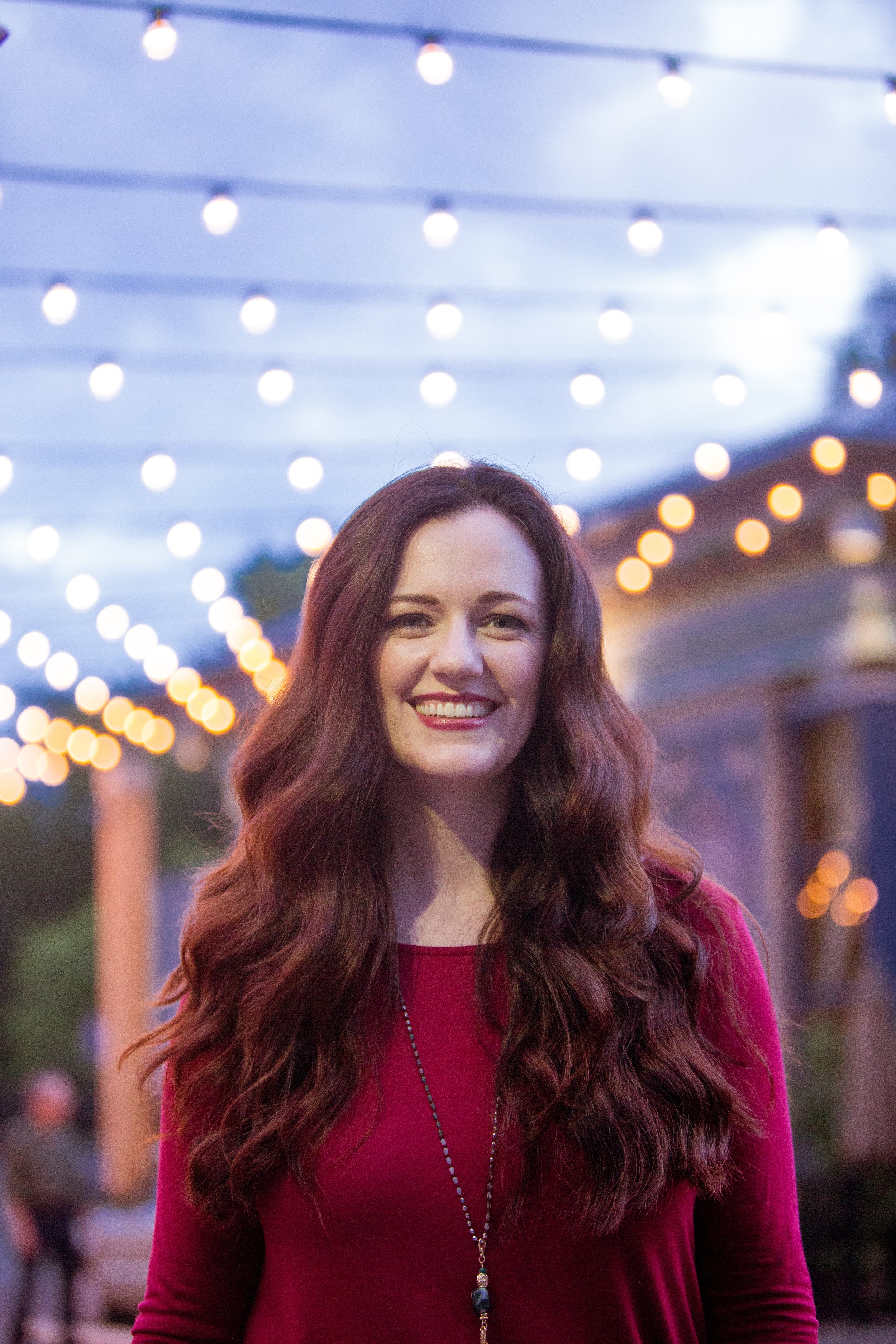 A woman with long, wavy red hair smiling outdoors at dusk, with string lights hanging overhead and blurred background.