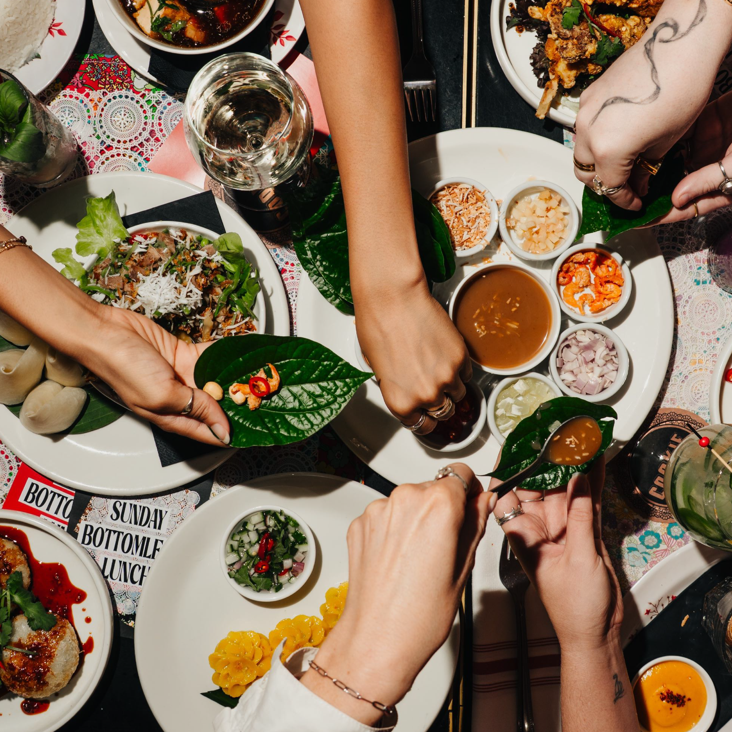 Top-down view of a lively Asian meal with hands serving and holding dishes, including a plate of salad with greens and shredded vegetables, a platter of small bowls with sauces and garnishes, a bowl of soup, and other dishes, on a table decorated with flowers and patterned tablecloths.