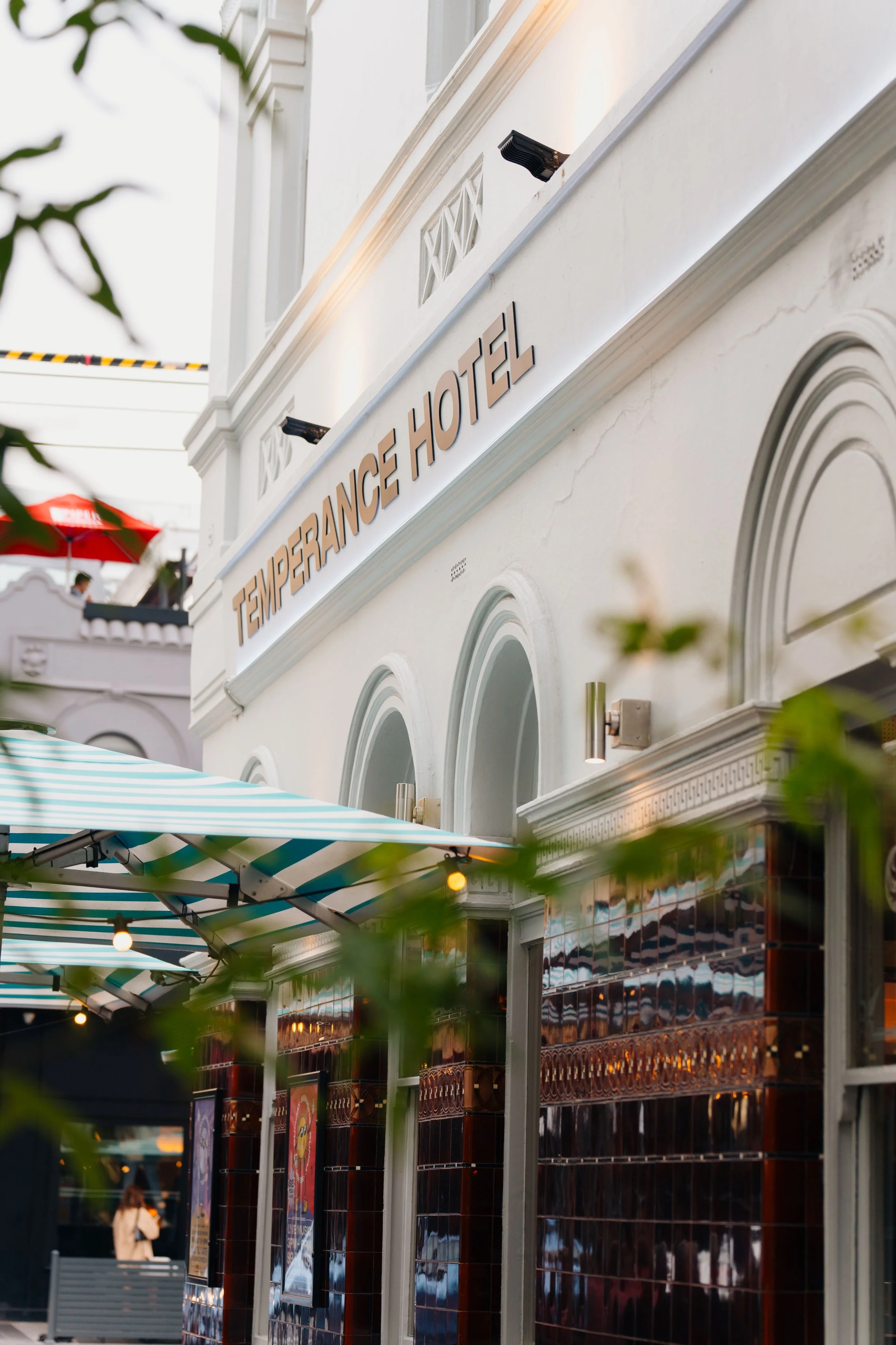 Exterior of the Temperance Hotel with a white facade, arched windows, and a sign displaying the hotel's name. There are outdoor striped awnings, greenery, and some posters on the wall.