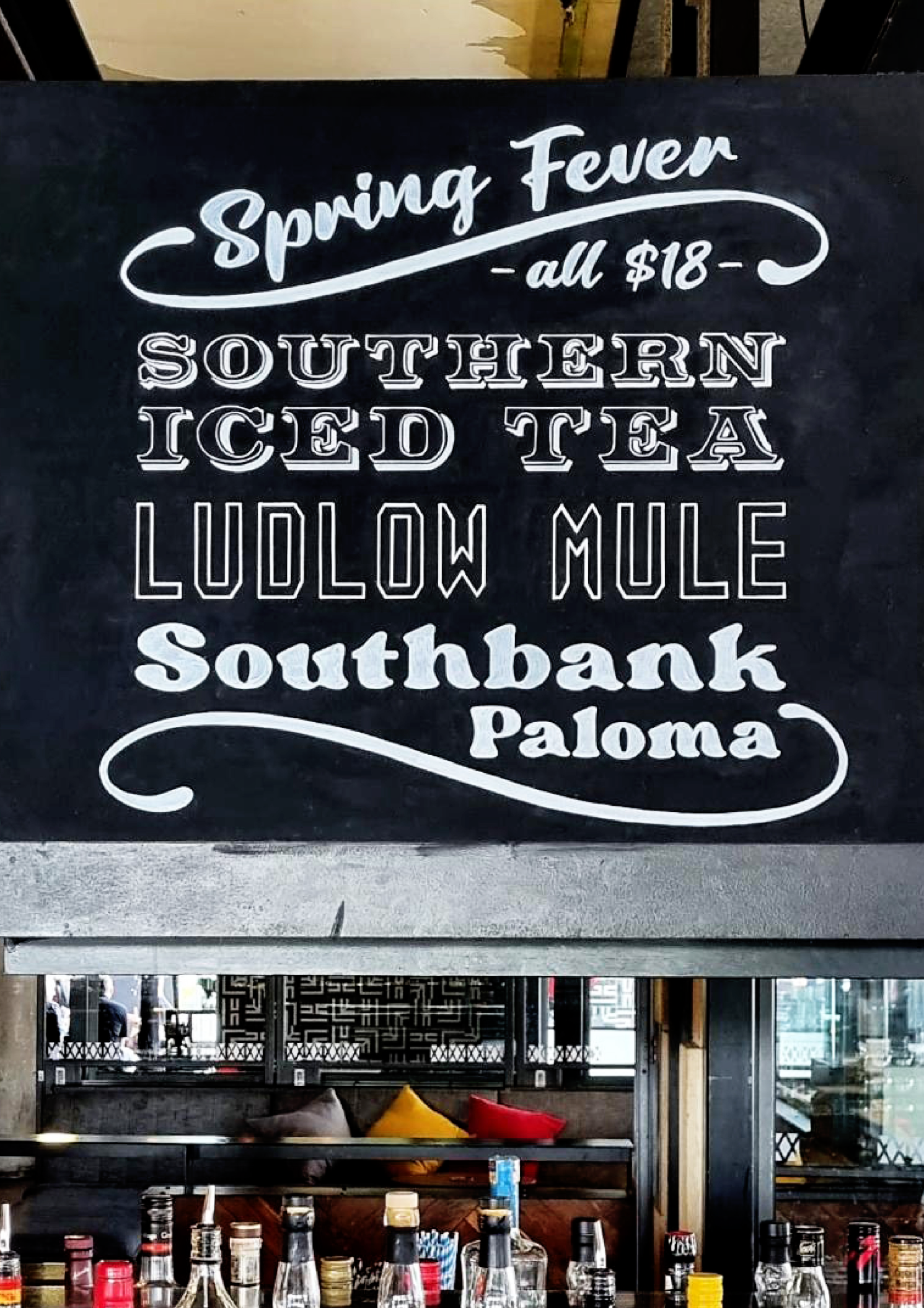 A chalkboard menu features a list of seasonal drinks priced at $18. The drinks include Southern Iced Tea, Ludlow Mule, Southbank, and Paloma. The setting appears to be a bar or restaurant with bottles and glasses on the counter and a seating area wit