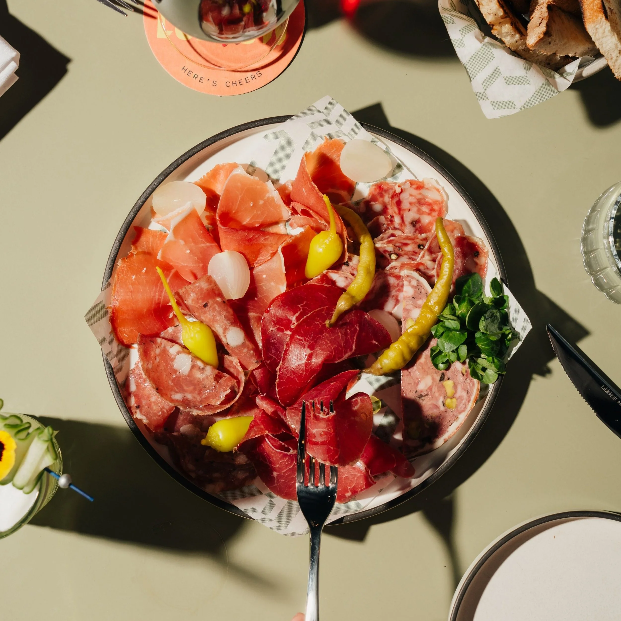 A plate of assorted cured meats garnished with small yellow peppers and parsley, placed on a light green table with other food items and a glass of wine nearby.