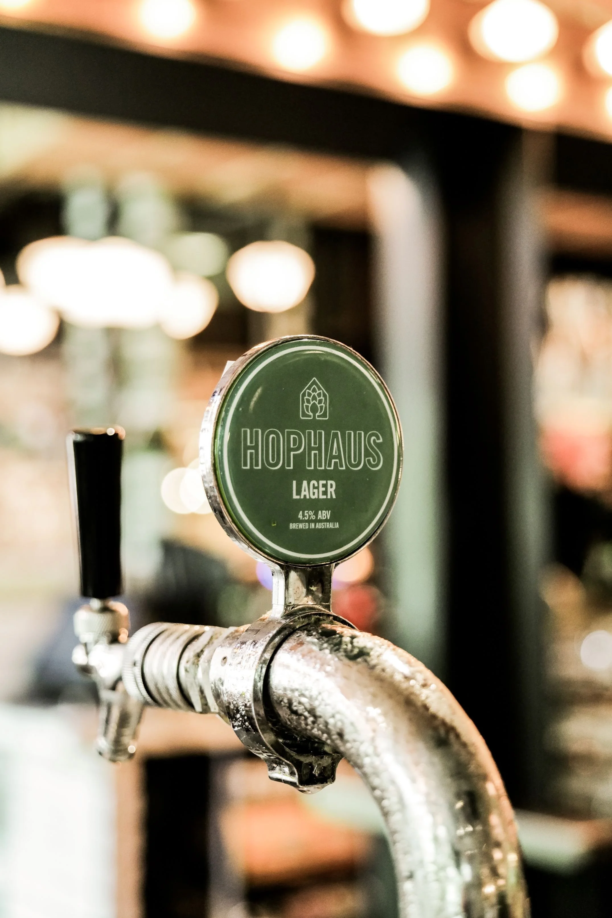 Close-up of a beer tap with a green sign labeled 'HOPHAUS Lager, 4.5% ABV, brewed in Australia' in a bar or pub setting.