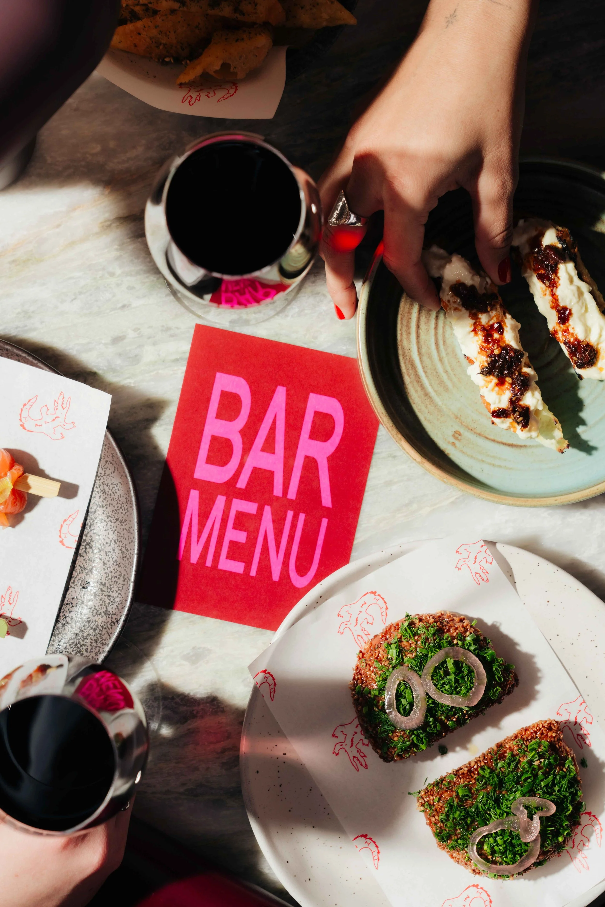 Overhead view of a bar table with a pink card labeled 'BAR MENU', two glasses of red wine, a black plate with three slices of cheesecake topped with fruit sauce, a white plate with two pieces of chopped meat garnished with onions, and a hand reaching for one of the cheesecakes.