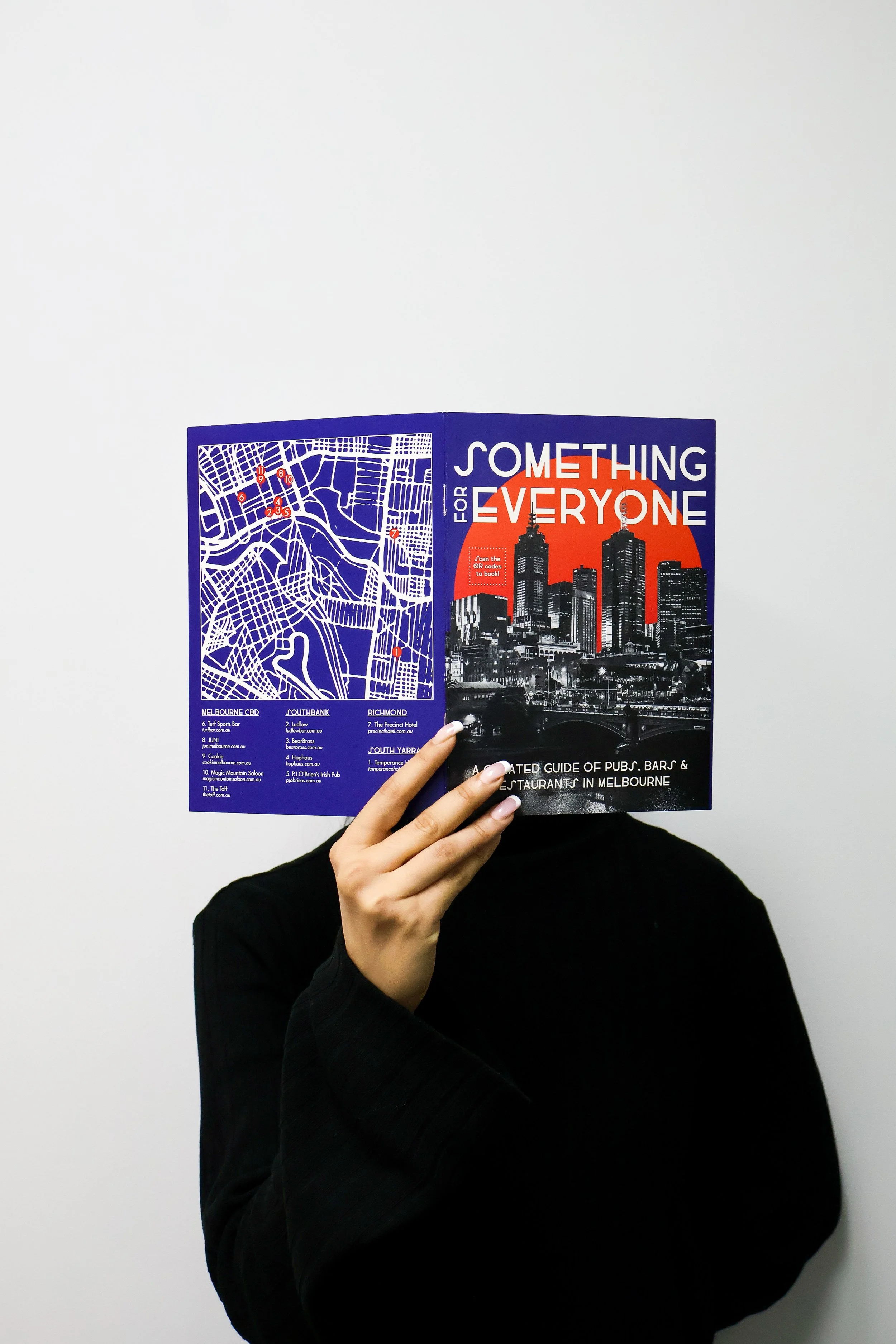 Person holding a travel guidebook covering their face, standing against a plain white wall.