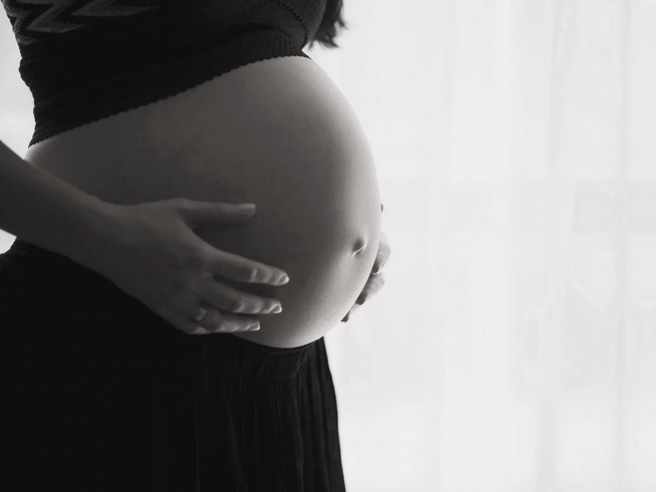 Black and white photo of a pregnant woman cradling her belly, standing near a light-colored curtain.