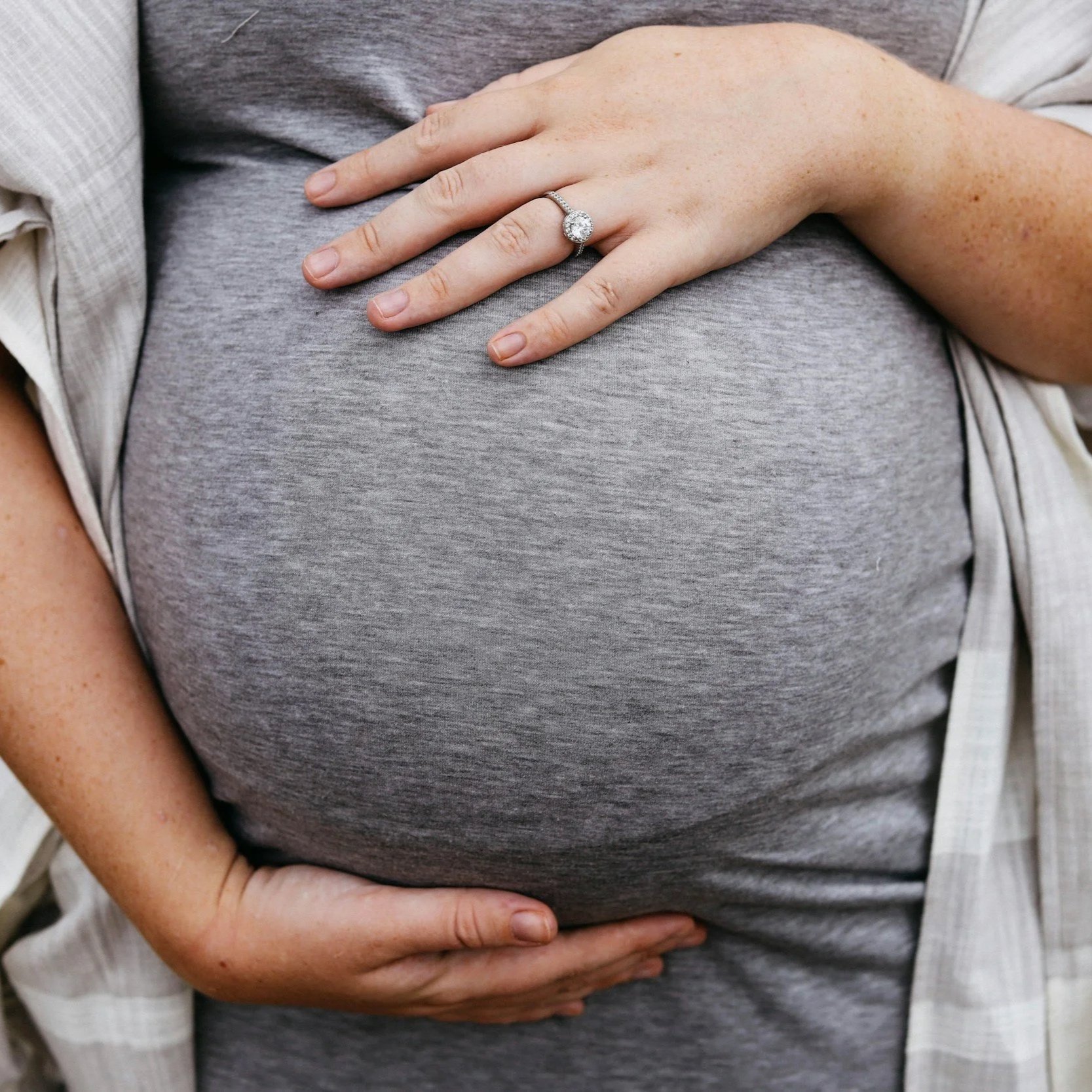 A pregnant belly with a woman’s hand resting on top, wearing an engagement ring.